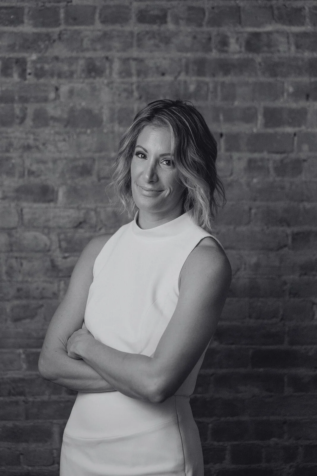 A black and white photo of a woman with wavy, shoulder-length hair, wearing a sleeveless white top, standing in front of a brick wall. She is smiling slightly with her arms crossed.