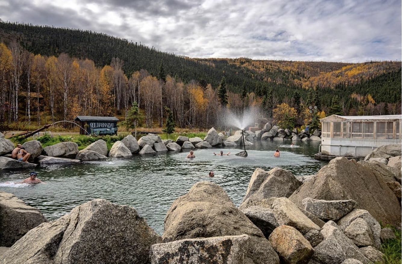 People relaxing and swimming in a natural hot spring surrounded by rocks in a mountainous area with trees and cloudy sky.