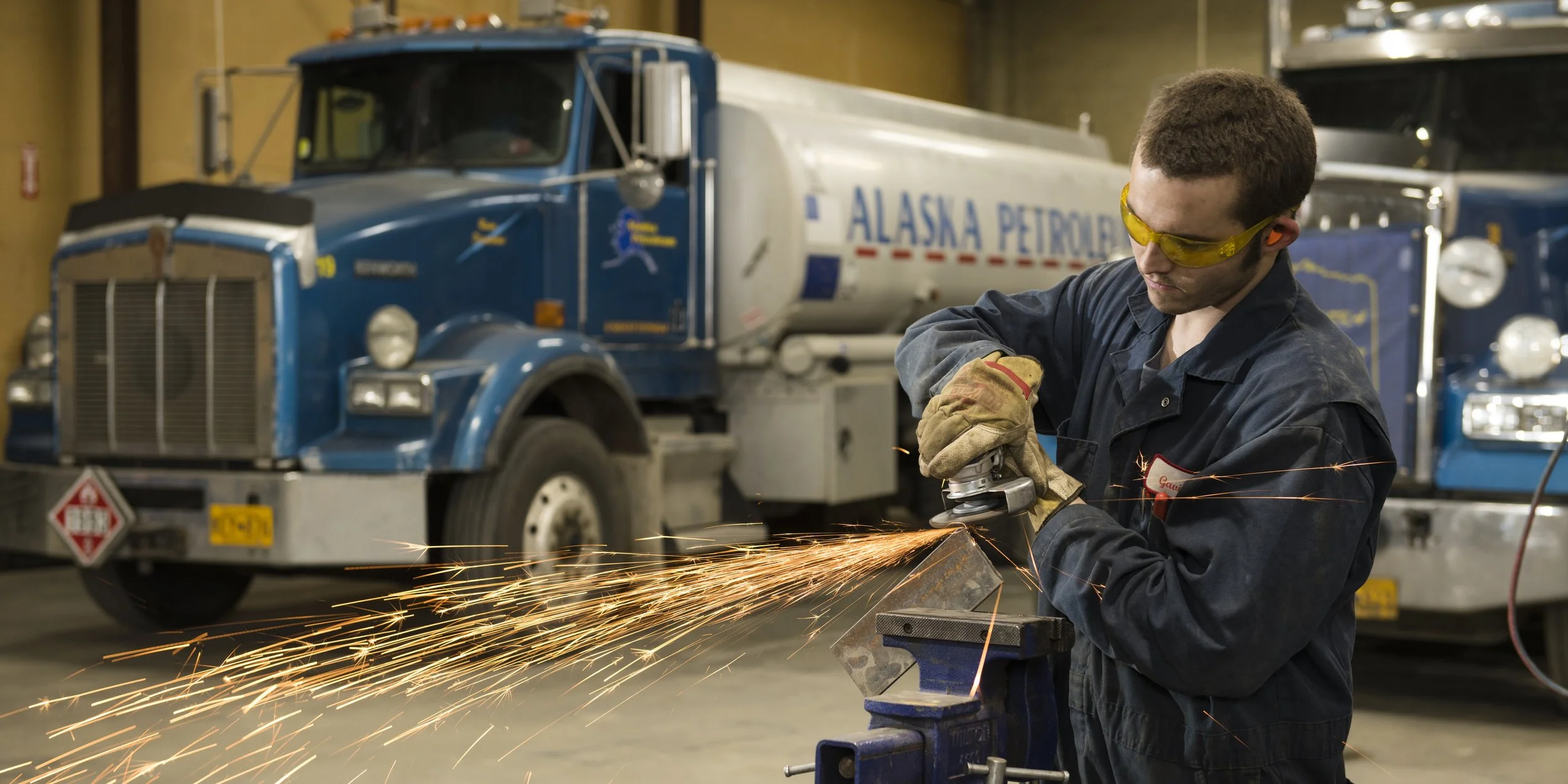 A worker wearing safety goggles and gloves uses a grinder on a metal piece, creating sparks. In the background, there are two blue trucks with 'Alaska Petroleum' written on them inside a garage or workshop.