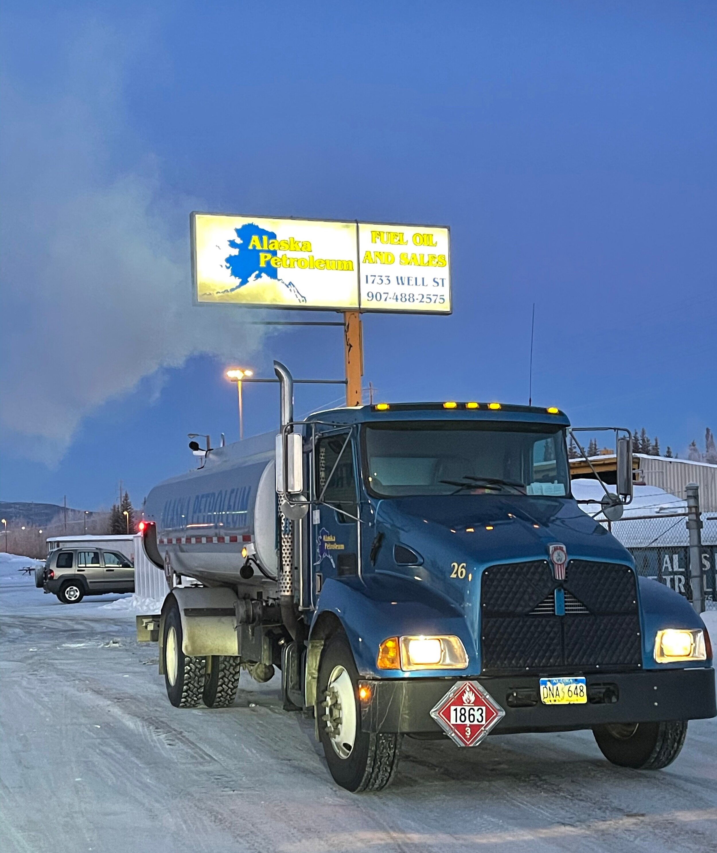A fuel truck parked in front of a sign that reads 'Alaska Petroleum, Fuel Oil and Sales, 1733 Well St, 907-488-2575'. The sign features a blue silhouette of Alaska. Snow on the ground, a fence, and in the background a few vehicles and a hill with sparse trees are visible.