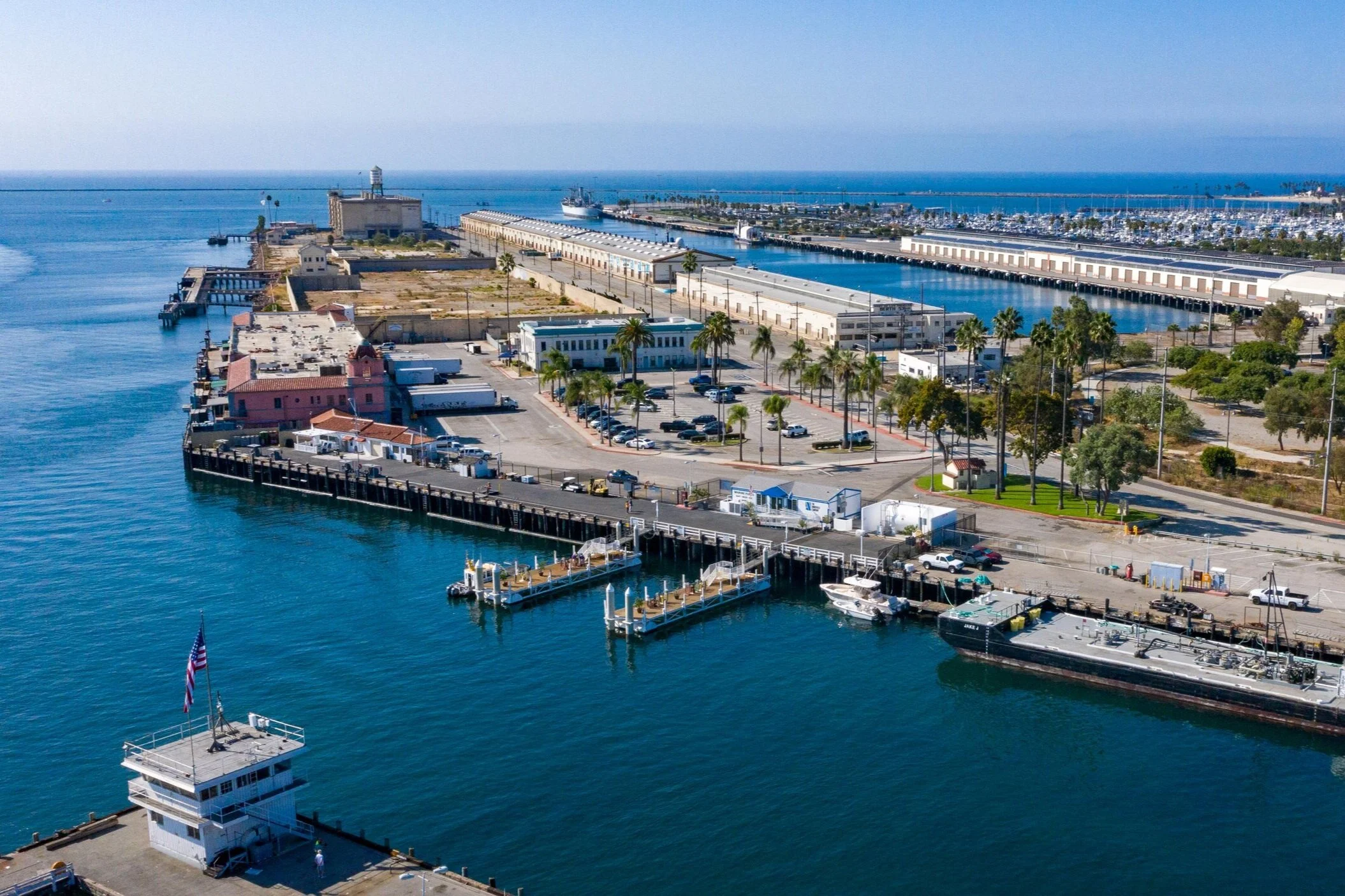 Aerial view of a harbor with docks, boats, and buildings, including an office tower, with a marina and the ocean in the background.