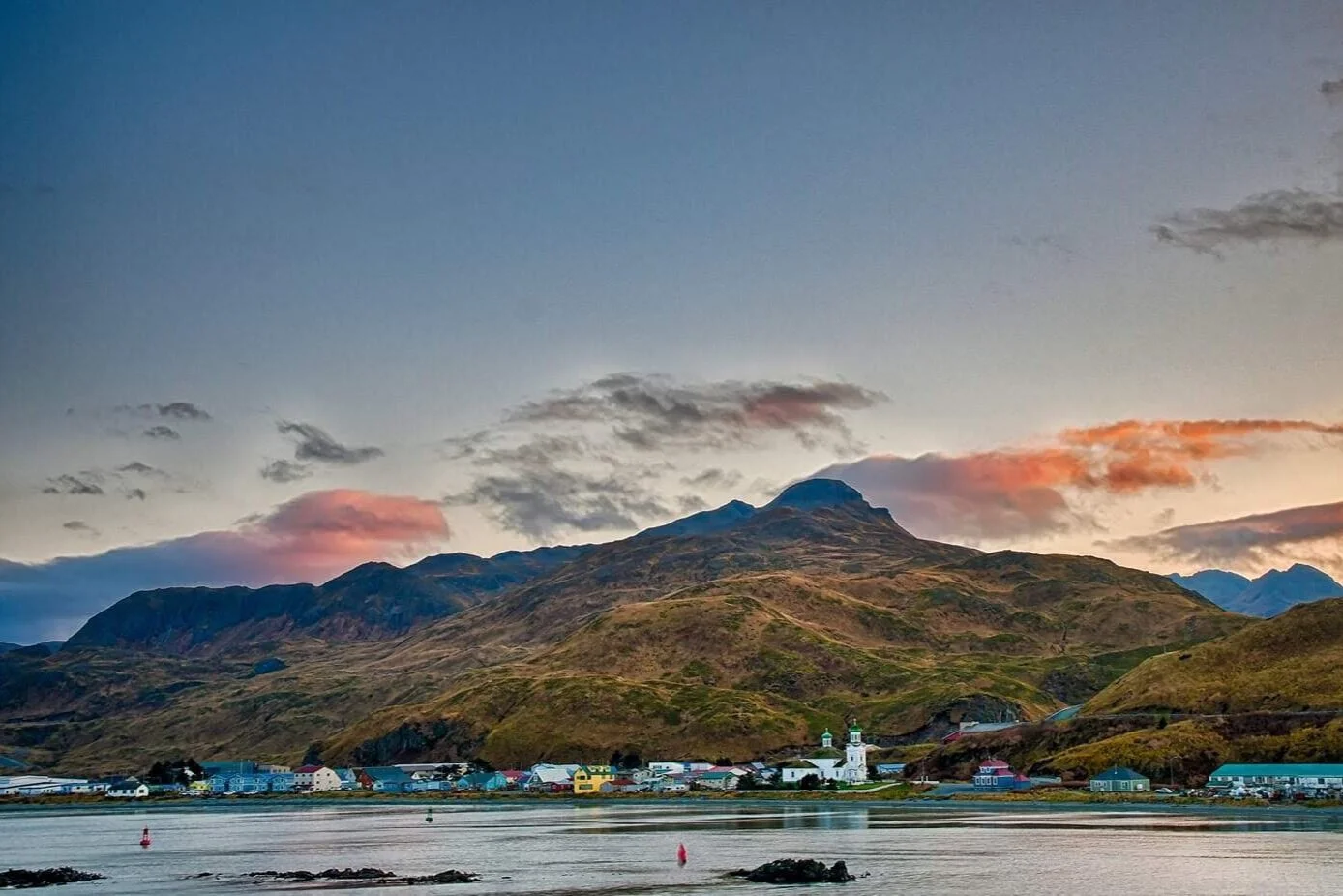 Scenic view of a coastal village at the foot of mountains during sunset, with calm water, colorful houses, a church, and a partly cloudy sky.