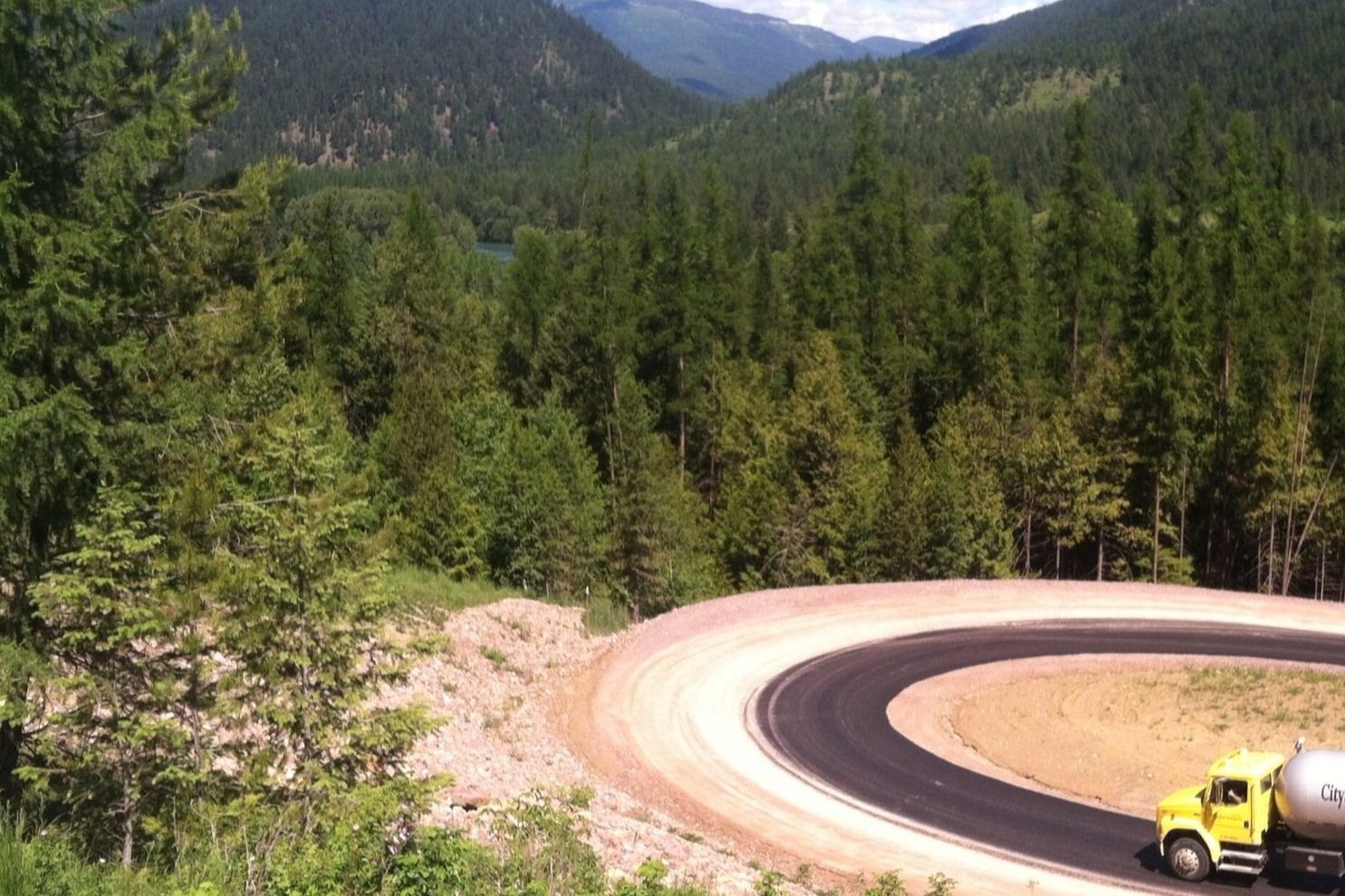 A yellow tanker truck driving on a winding mountain road surrounded by dense green pine trees, with distant mountains in the background.