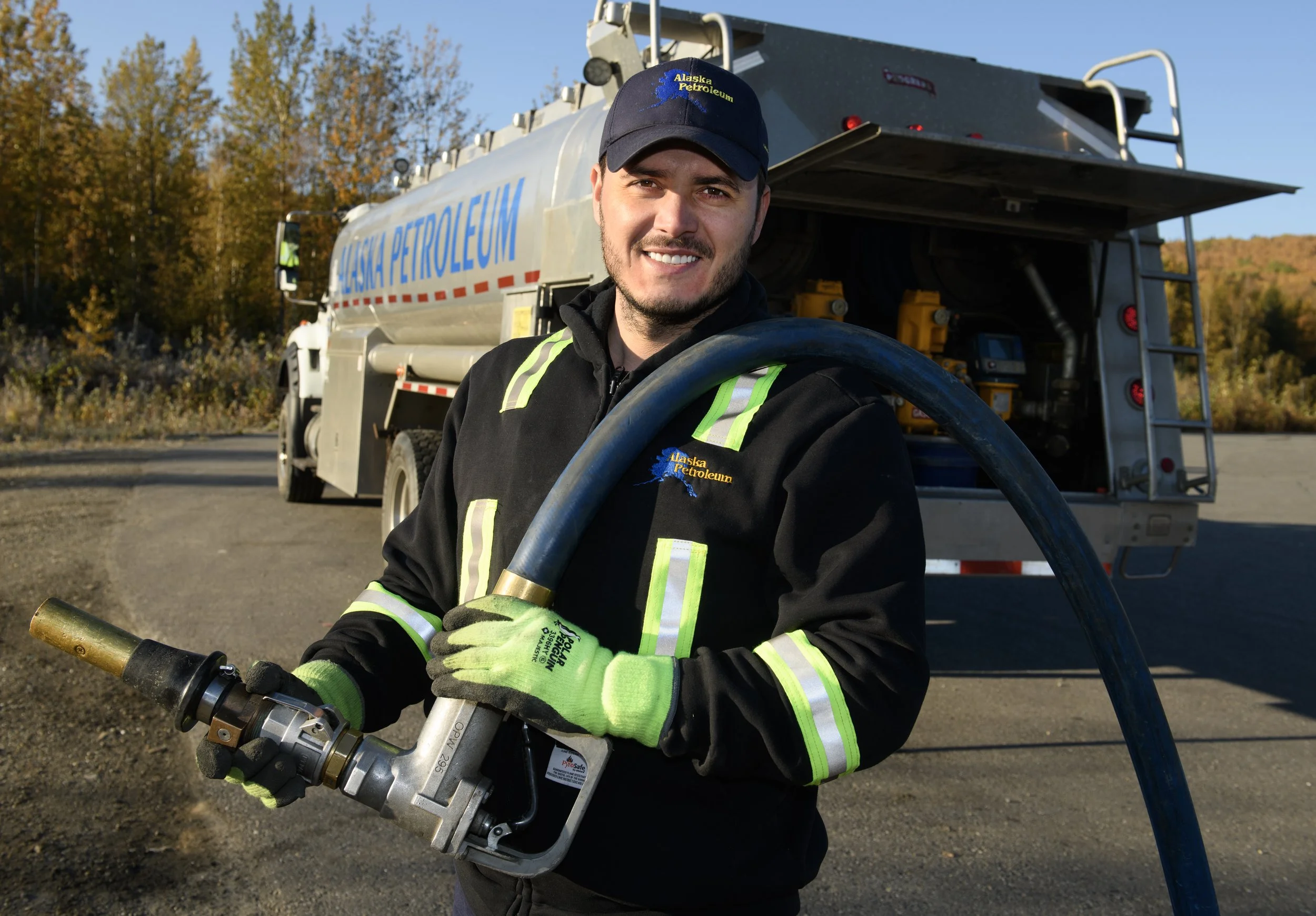 A man wearing a black uniform and neon green gloves holding a large pipe, standing in front of an Alaska Petroleum fuel truck, with a background of trees and a clear blue sky.