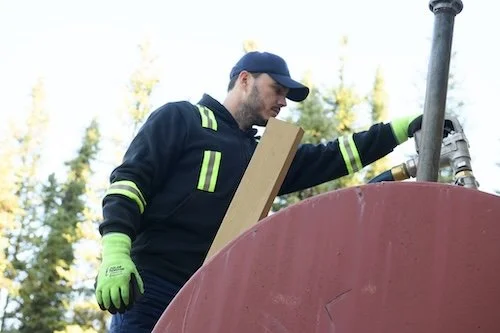 Man wearing a navy uniform and green gloves working outdoors, handling a metal pipe near a red tank or container.