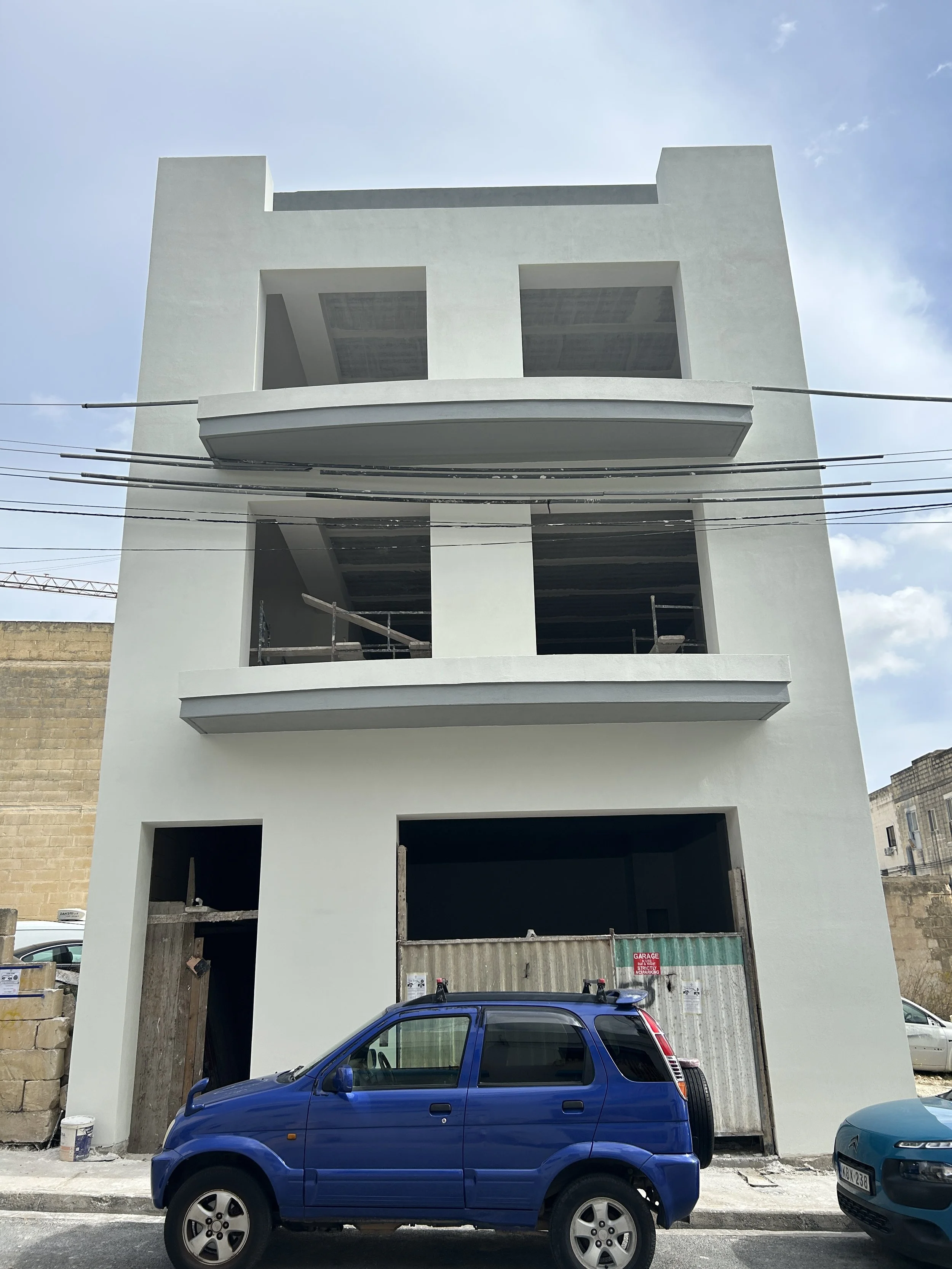 Four-story building under construction with blue car parked in front, construction materials visible, and blue sky overhead.