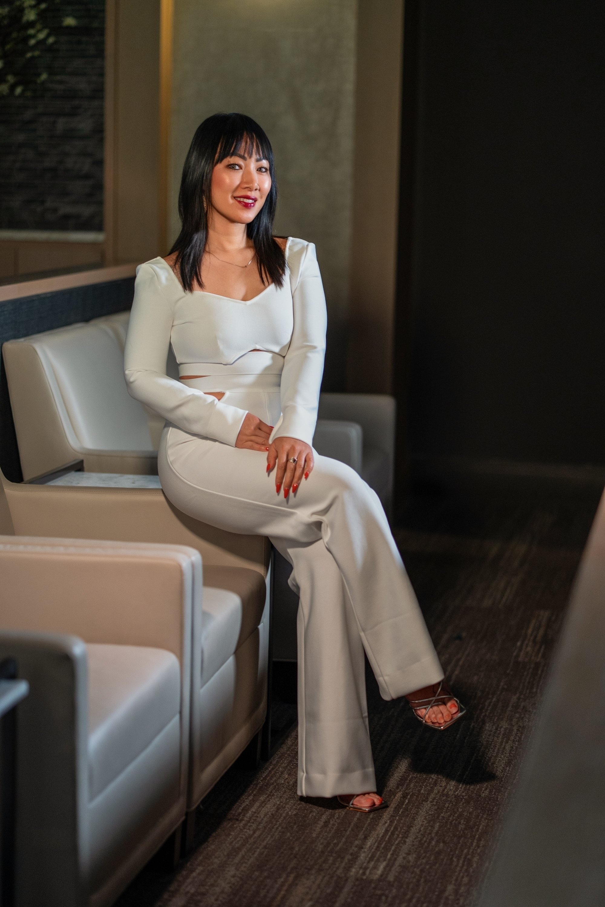 A woman sitting on a beige bench in an indoor setting, wearing a white pantsuit with cutouts, red lipstick, and high-heeled sandals, smiling and looking at the camera.
