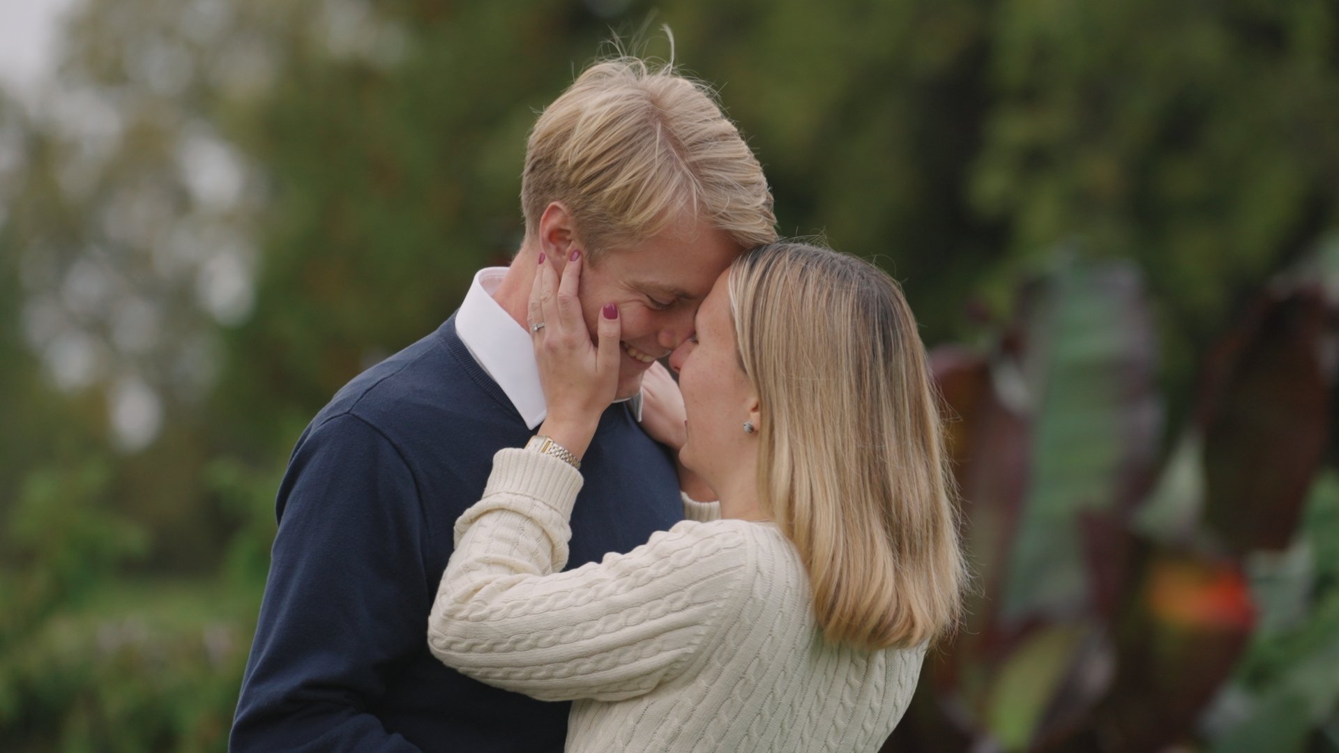 A couple sharing an intimate moment outdoors, touching foreheads and smiling, surrounded by blurred green and brown foliage.