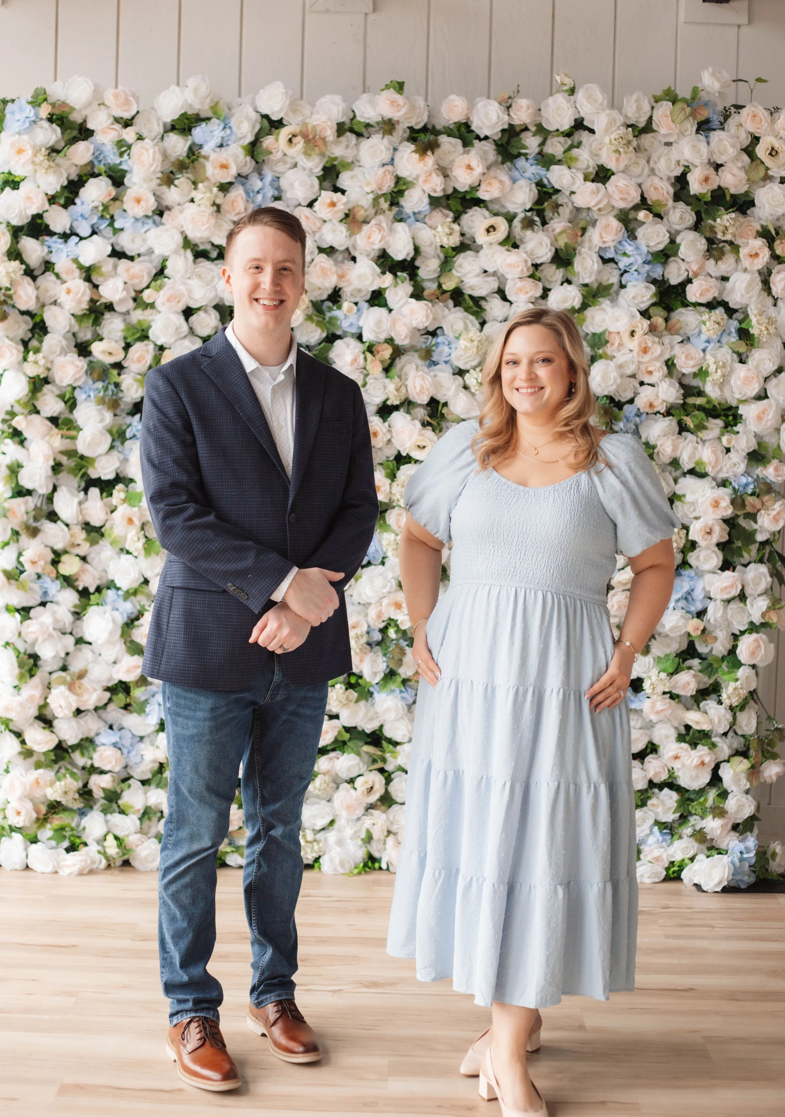 A man and a woman standing in front of a floral backdrop of white and light pink roses with greenery, smiling at the camera.