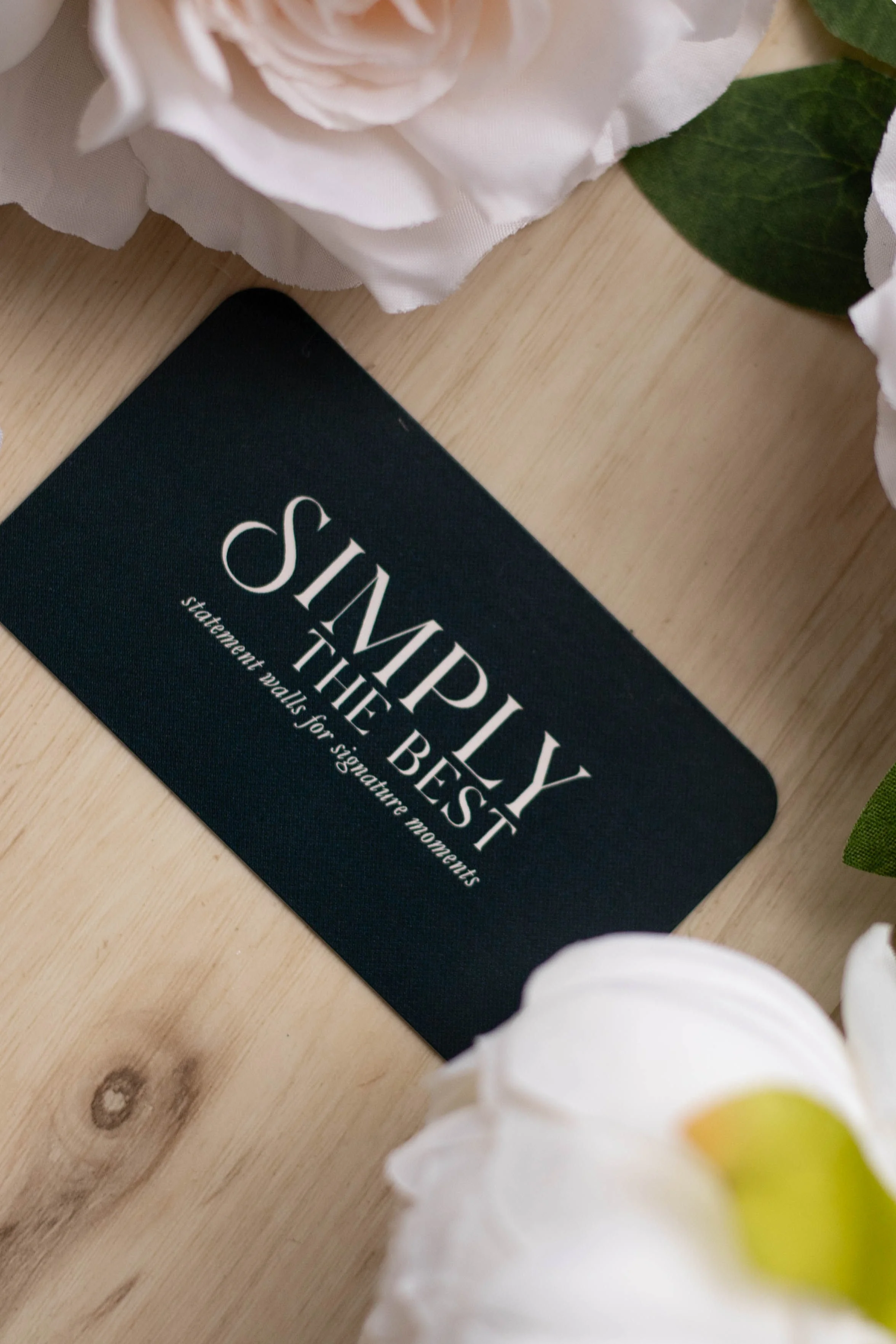 A black card with white text that says, 'SIMPLY THE BEST statement walls for signature moments,' placed on a light wooden surface, partially surrounded by white flowers and green leaves.