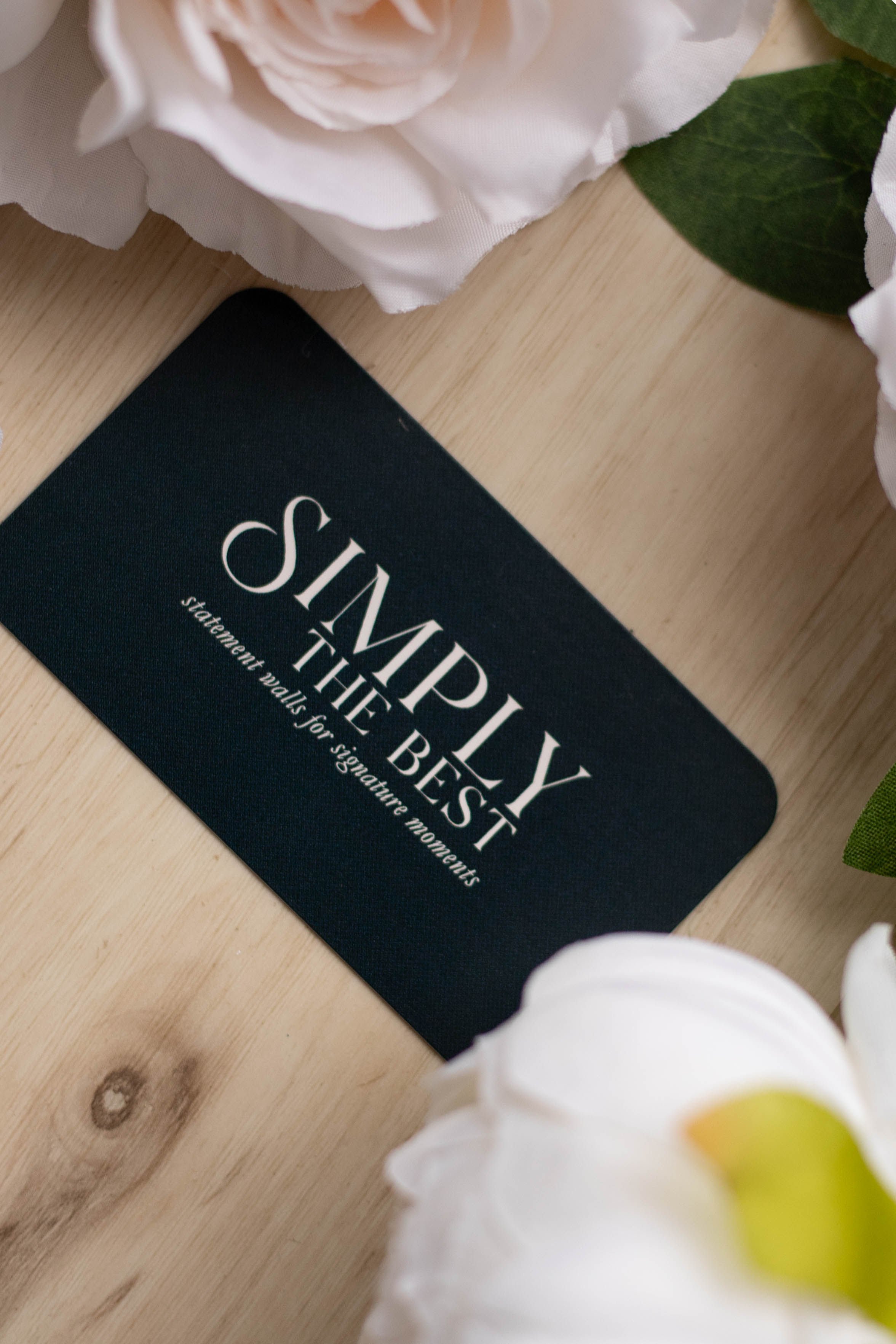 A black card with white text that says, 'SIMPLY THE BEST statement walls for signature moments,' placed on a light wooden surface, partially surrounded by white flowers and green leaves.