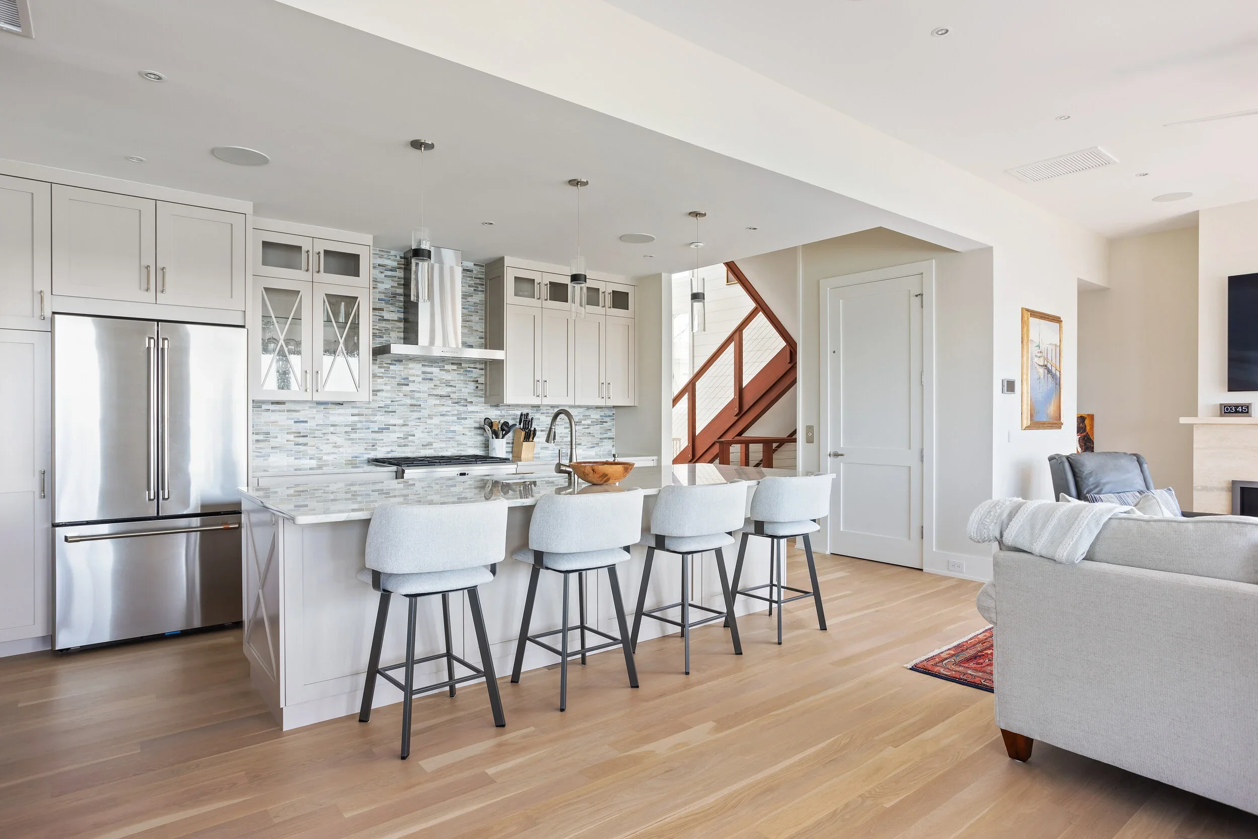Modern open-concept kitchen with white cabinets, a marble backsplash, stainless steel appliances, and a breakfast bar with four barstools, adjacent to a living room with a gray sofa and a staircase with wooden railing.