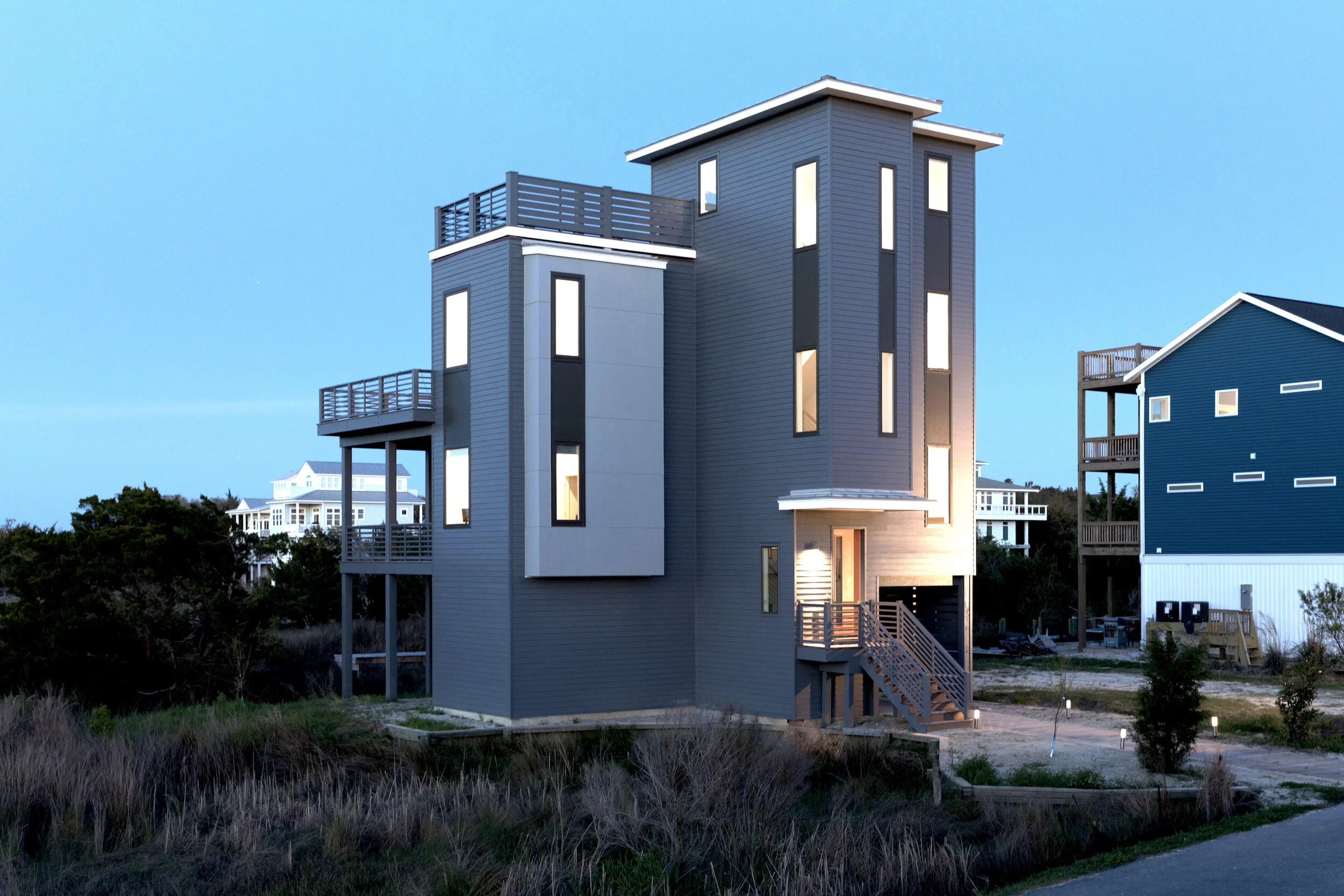 A modern, multi-story house with narrow vertical windows, multiple balconies, and dark gray siding, situated in a coastal area during dusk.