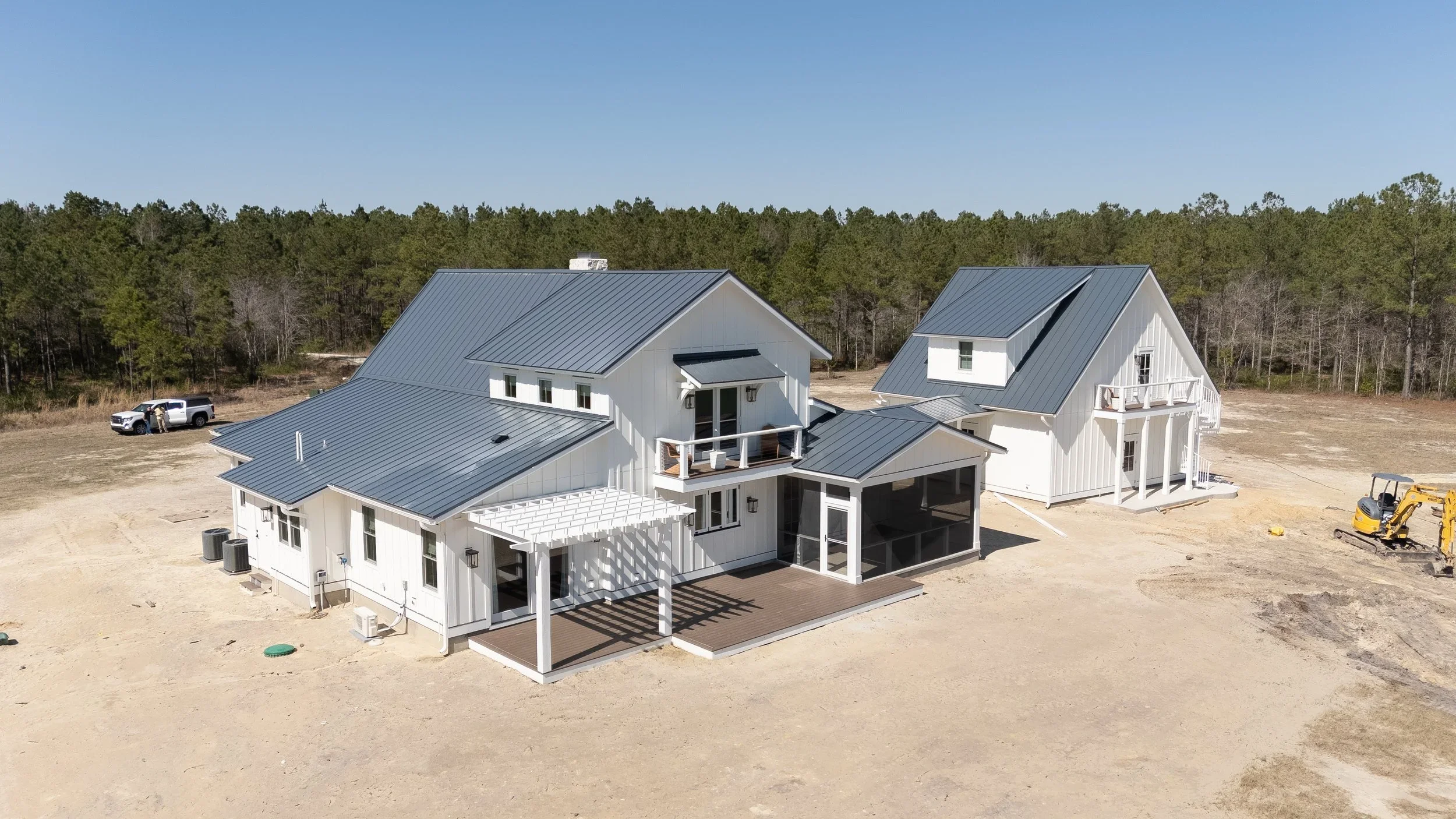 Aerial view of two white modern houses with metal roofs on a large plot of land under construction, surrounded by trees in the background.