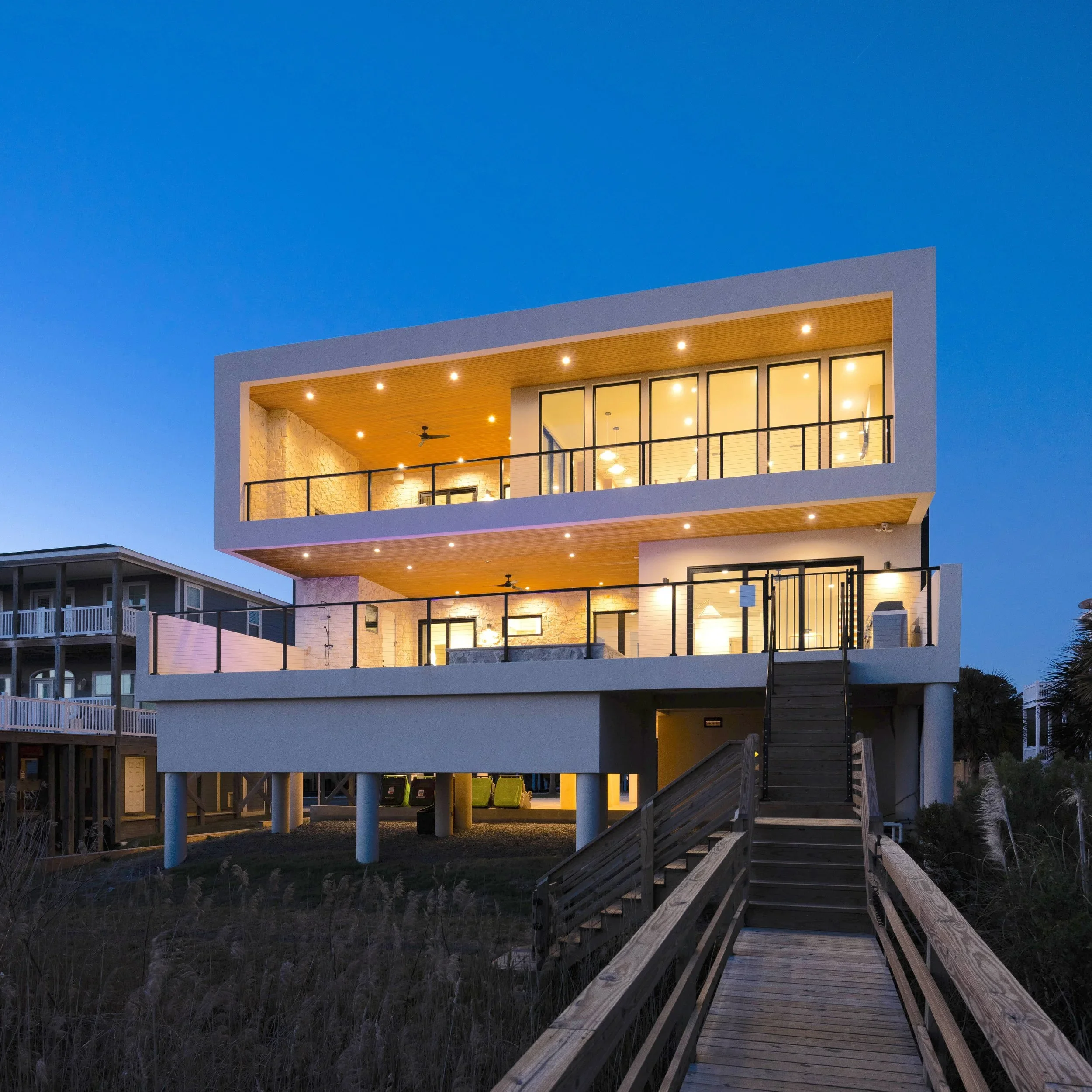Modern three-story house with large glass windows and balconies, built on stilts, illuminated at dusk, with a wooden ramp leading to the entrance.