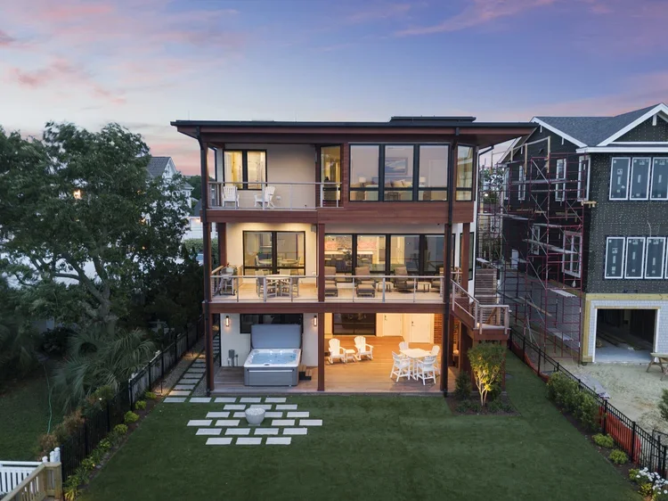Modern three-story house with large glass windows, outdoor seating, and a backyard with a hot tub and landscaped lawn during sunset.