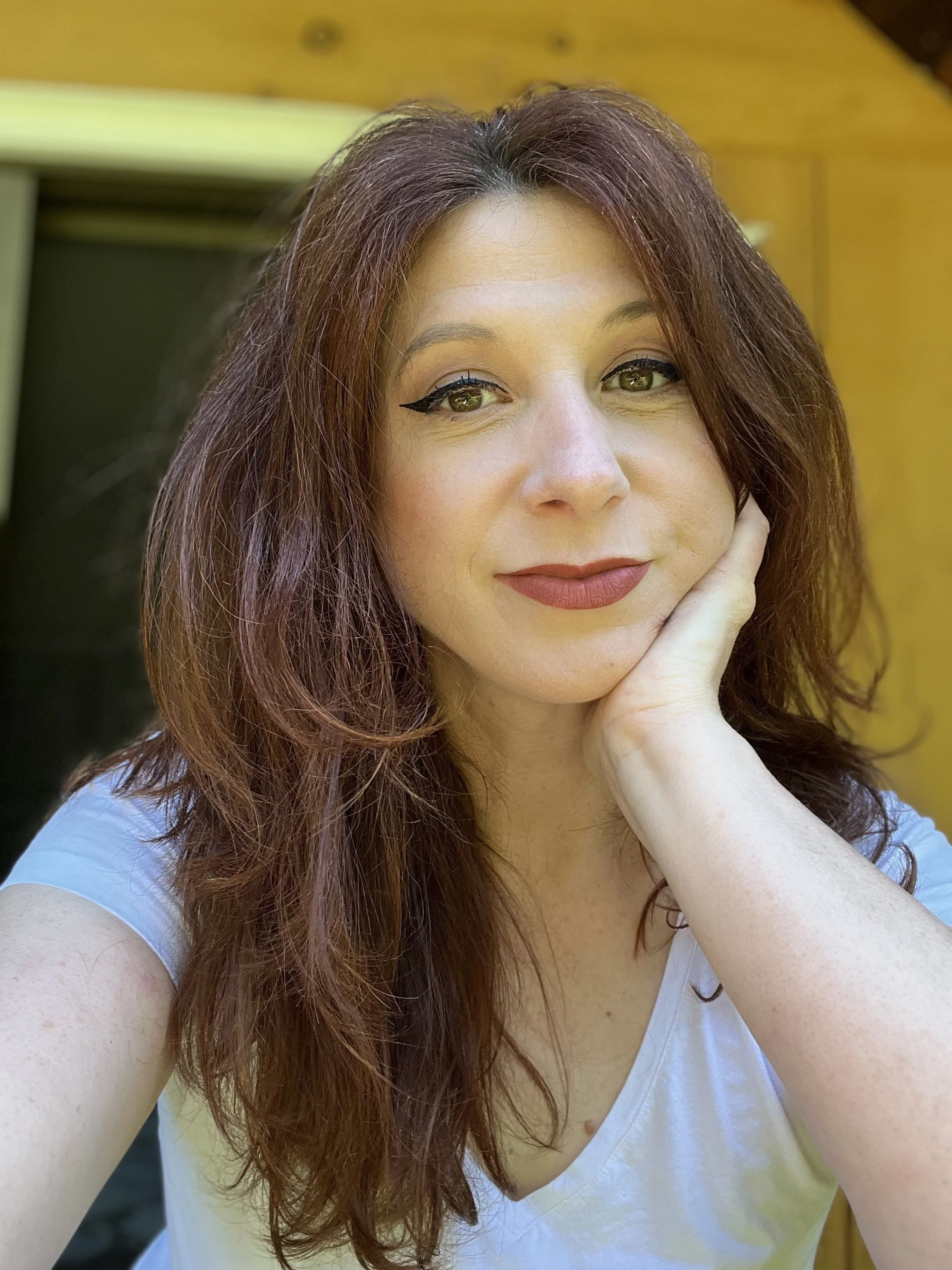 A woman with red hair resting her face on her hand, wearing a white top, with a wooden wall in the background.