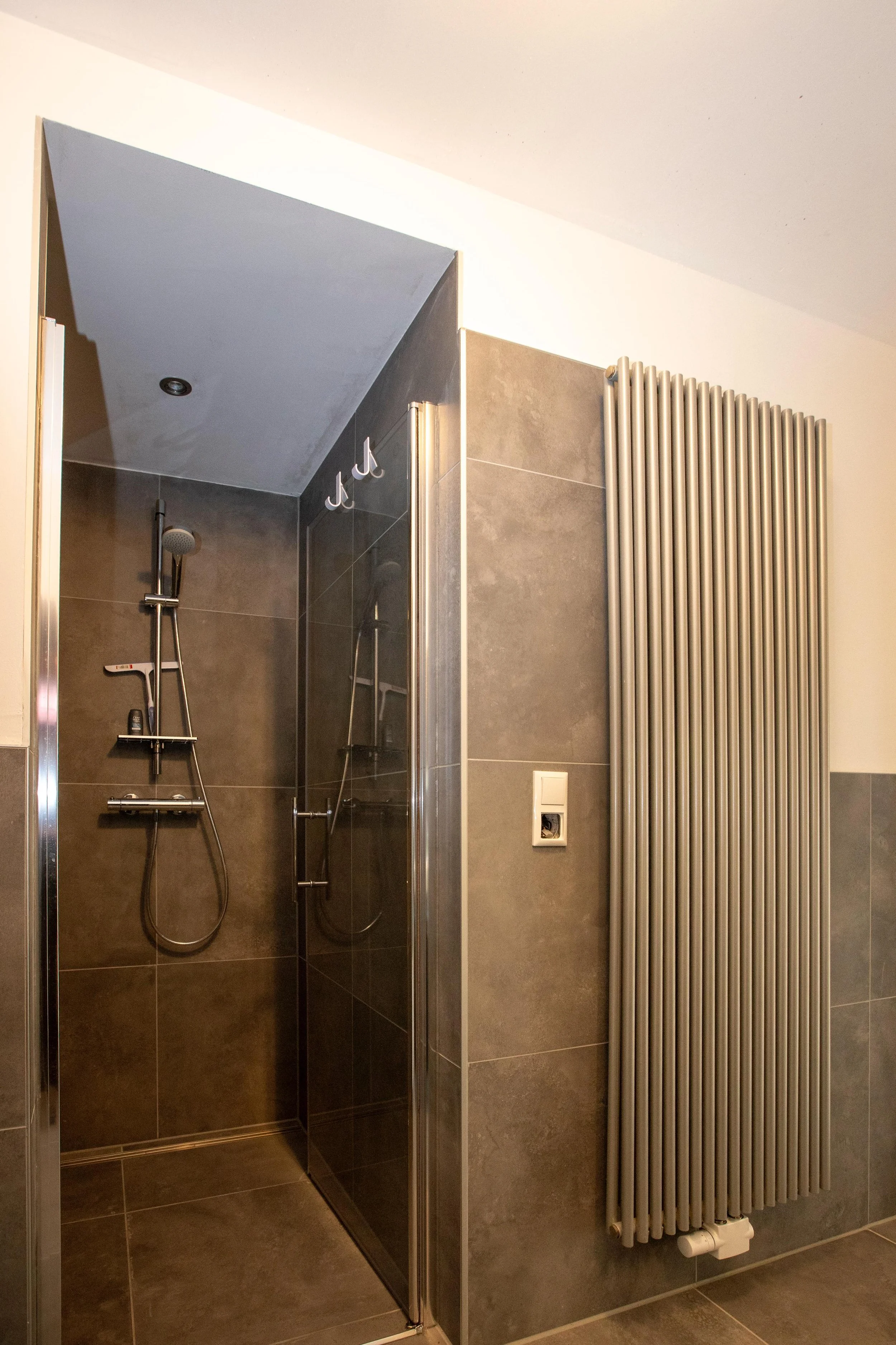Dusche mit grauen Fliesen, Duschkopf und Handbrause, neben einem großen Funkkörperheizkörper an der Wand.