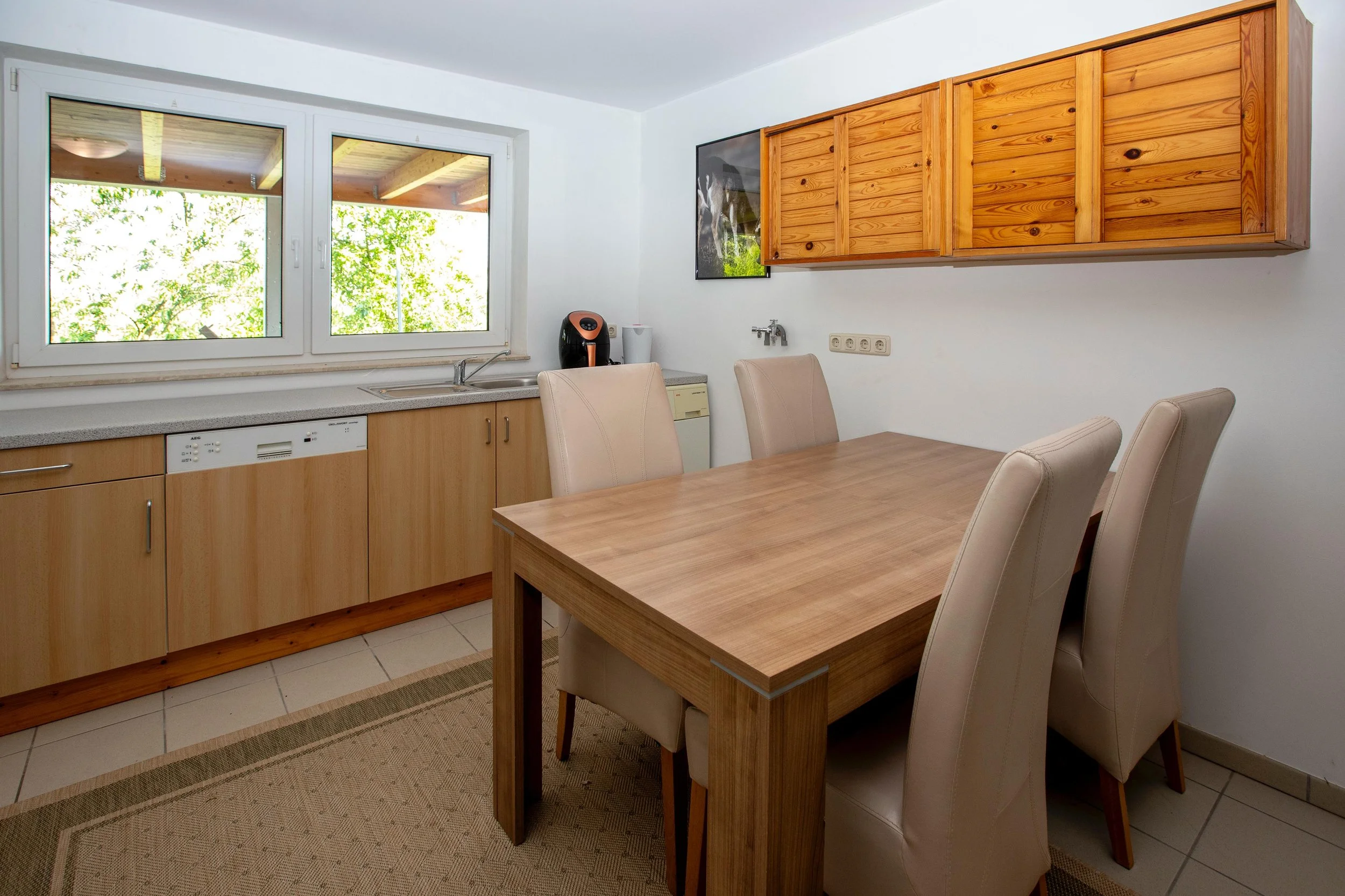 Küchenraum mit Holzmöbeln, Couchtisch, weißen Ledersesseln, Fenster mit Blick auf grüne Bäume, Kaffeeautomat und Geschirrspüler.