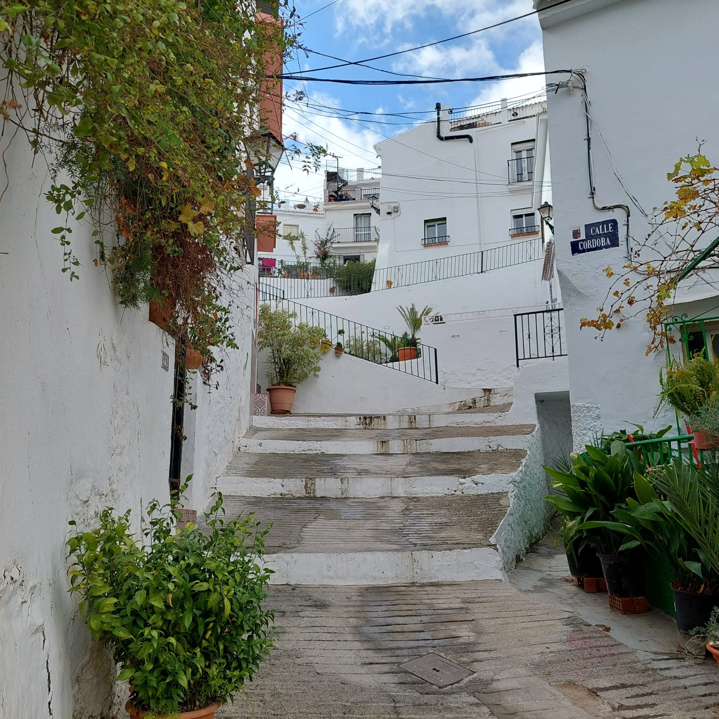 White-washed narrow street in a hilltown with potted plants and stairs, indicating a Mediterranean or Andalusian setting, with a street sign saying 'Calle Cordoba'.