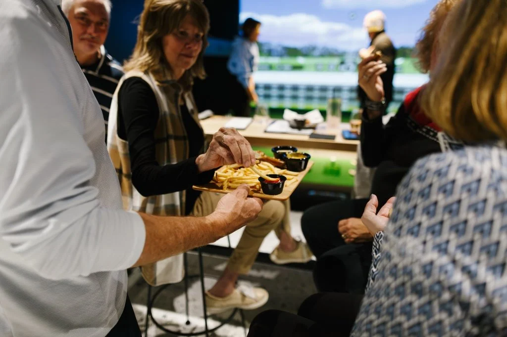 People enjoying food and drinks at a social gathering, with a woman being served fries and dipping sauces at a table with others, and a background of a window showing a bright day outside.