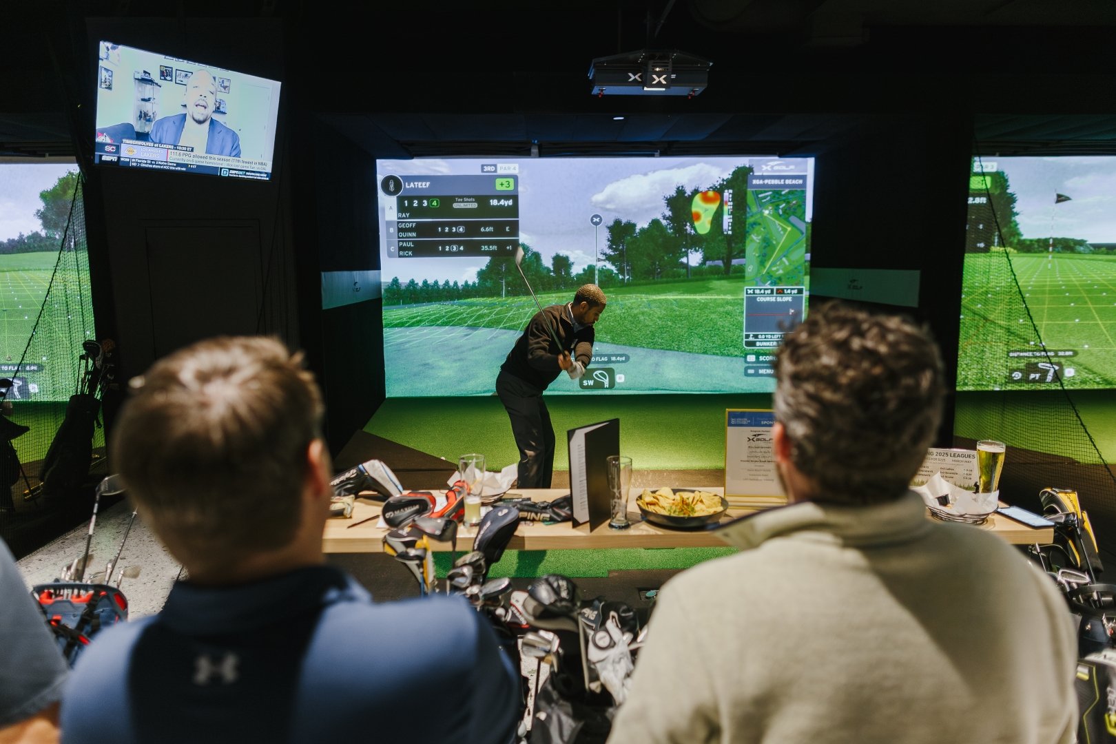 Two men sitting at a golf simulation center watching a man in a black jacket swinging a golf club on a virtual golf course displayed on large screens.