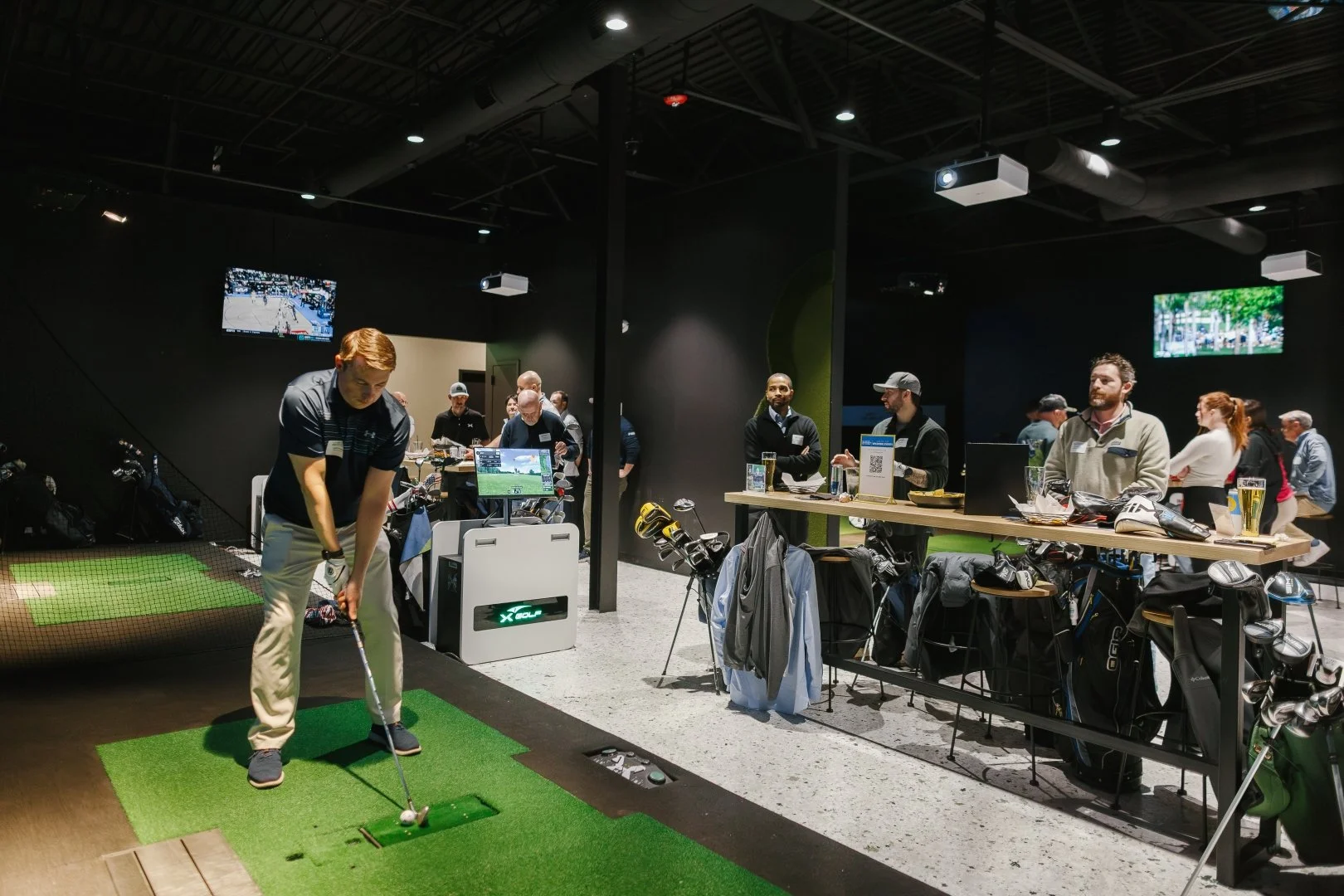 Person practicing golf at an indoor golf simulator, with others observing and socializing nearby.
