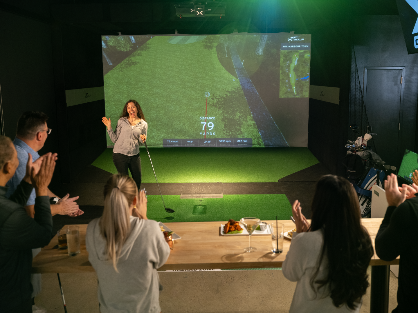 A group of people in a golf simulation room watching a woman prepare to hit a golf ball. The woman stands in front of a large screen displaying a golf course with distance and swing data. Some audience members are clapping and smiling.