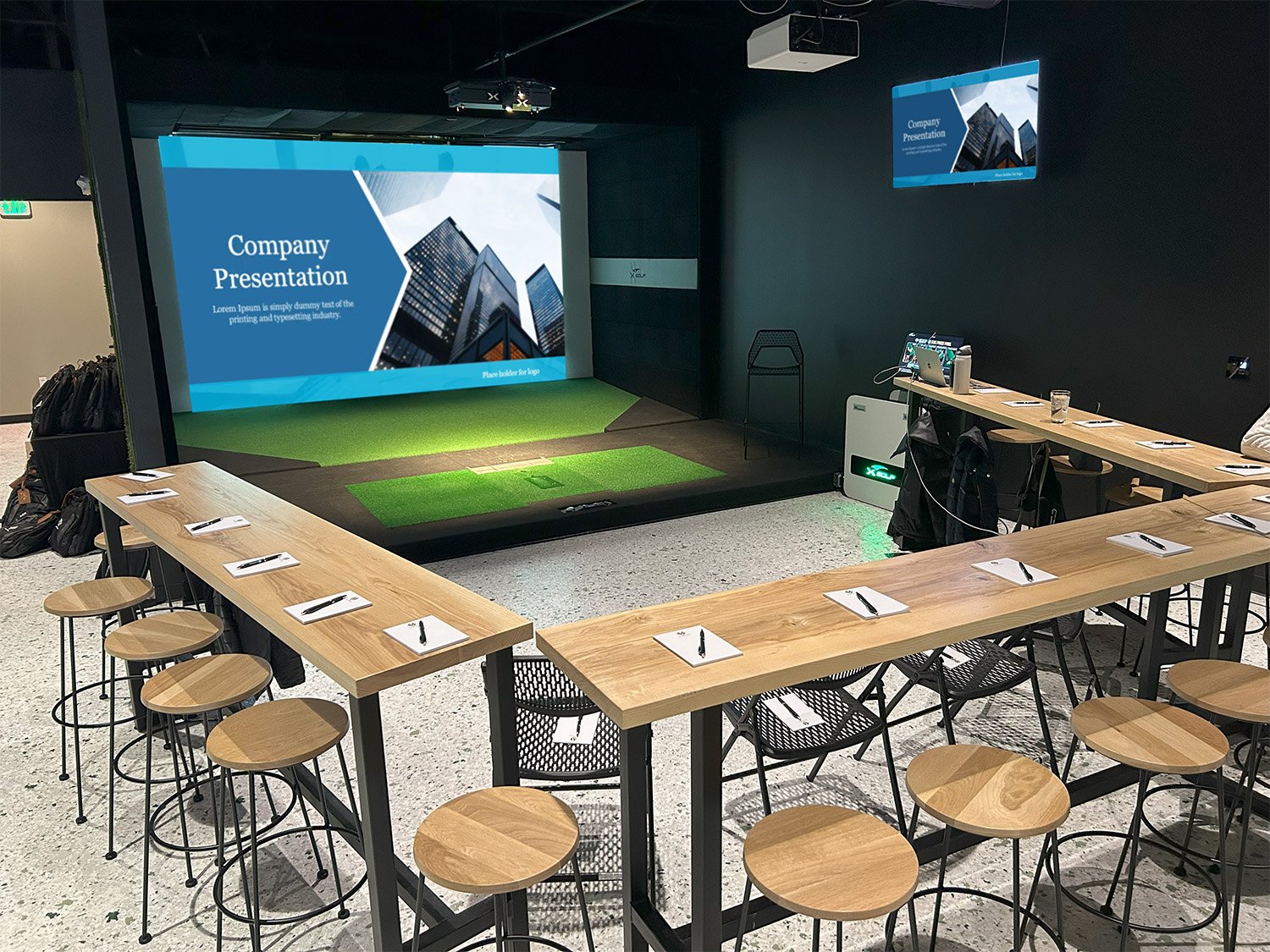 A conference room set up for a presentation with a U-shaped table arrangement, notebooks and pens at each seat, a small stage with a large projected screen displaying a cityscape and presentation slide, and a smaller screen on the wall showing the same slide.