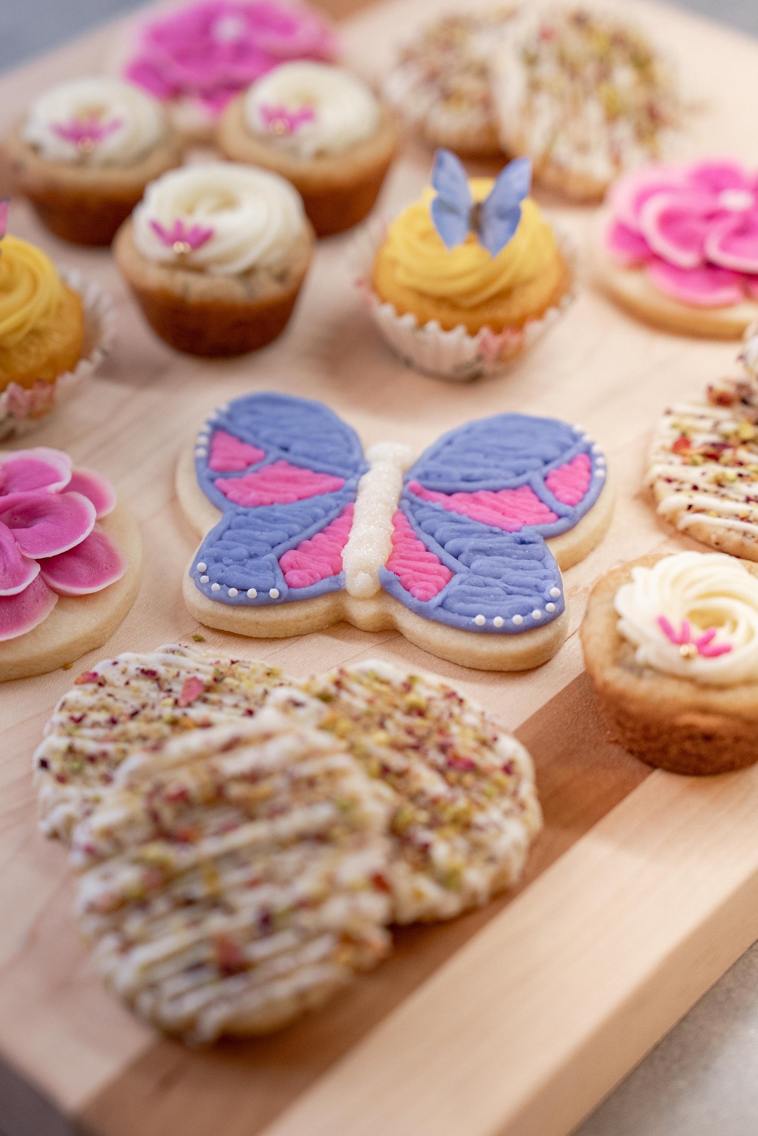 A buttercream frosted sugar cookie in the shape of a butterfly is surrounded by cookie cups, cupcakes and shortbread cookies on a light wood cutting board