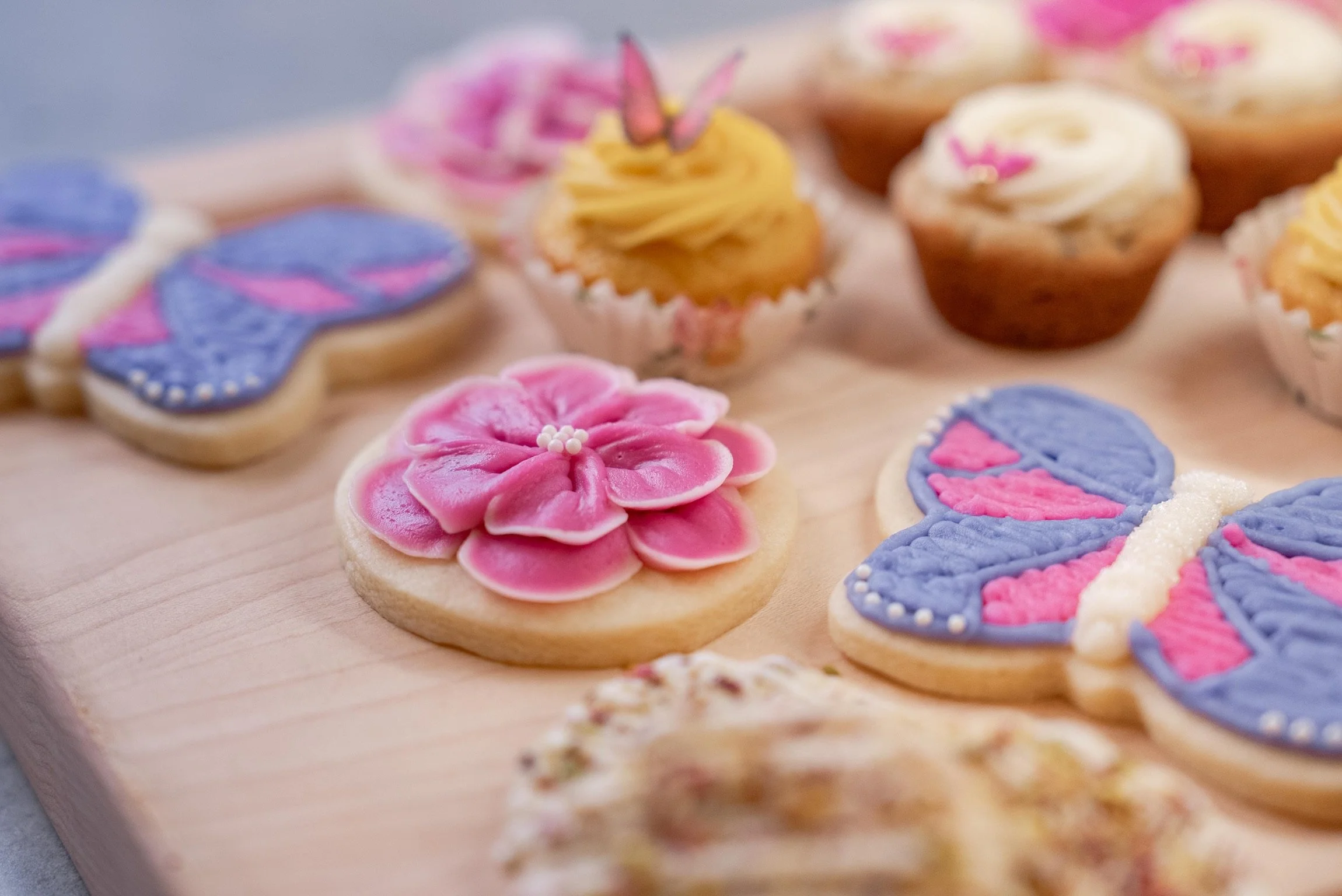 Colorful decorated cookies and cupcakes on a wooden surface, including a pink flower-shaped cookie with white sugar pearl center and butterfly-shaped cookies with purple and pink icing.