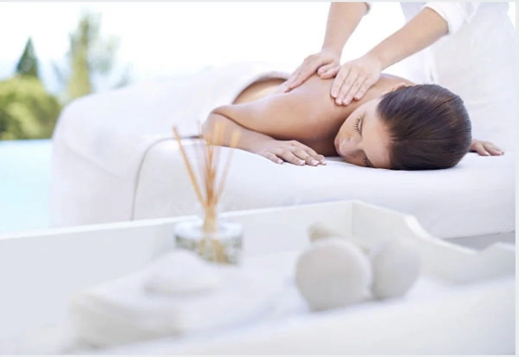 A woman receiving a back massage on a white massage table outdoors with greenery in the background.