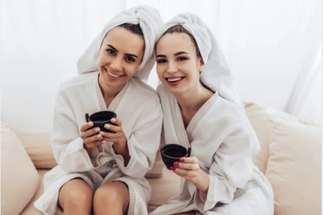 Two women with towels on their heads and robes, sitting on a bed, smiling and holding coffee cups in a bright, cozy room.