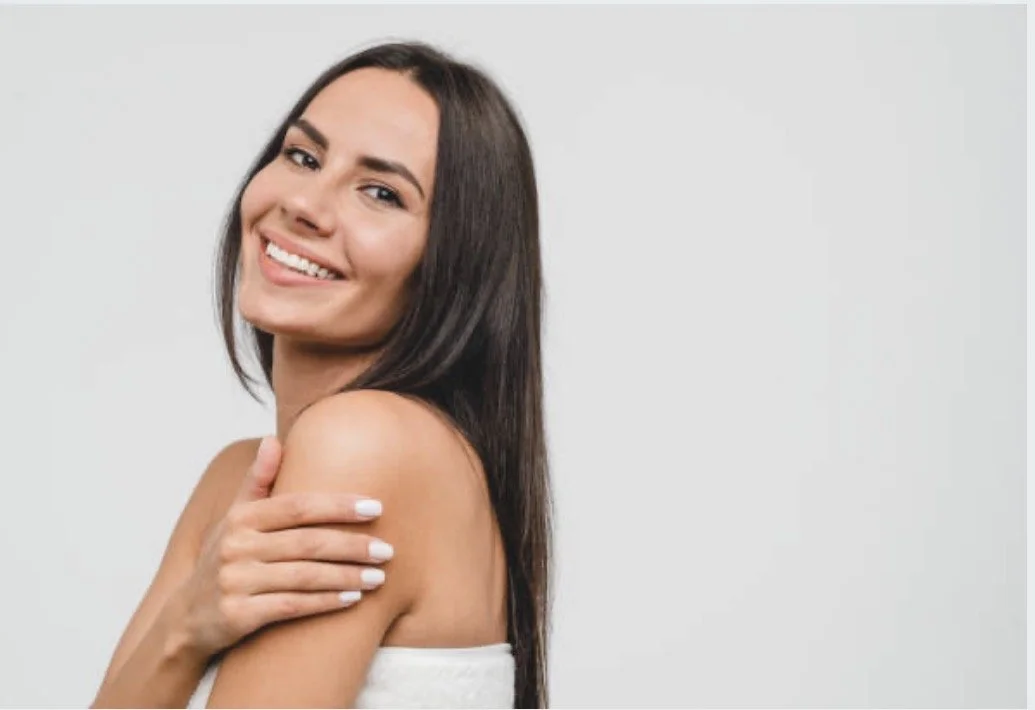 A young woman with long brown hair smiling and touching her shoulder, standing against a plain white background.