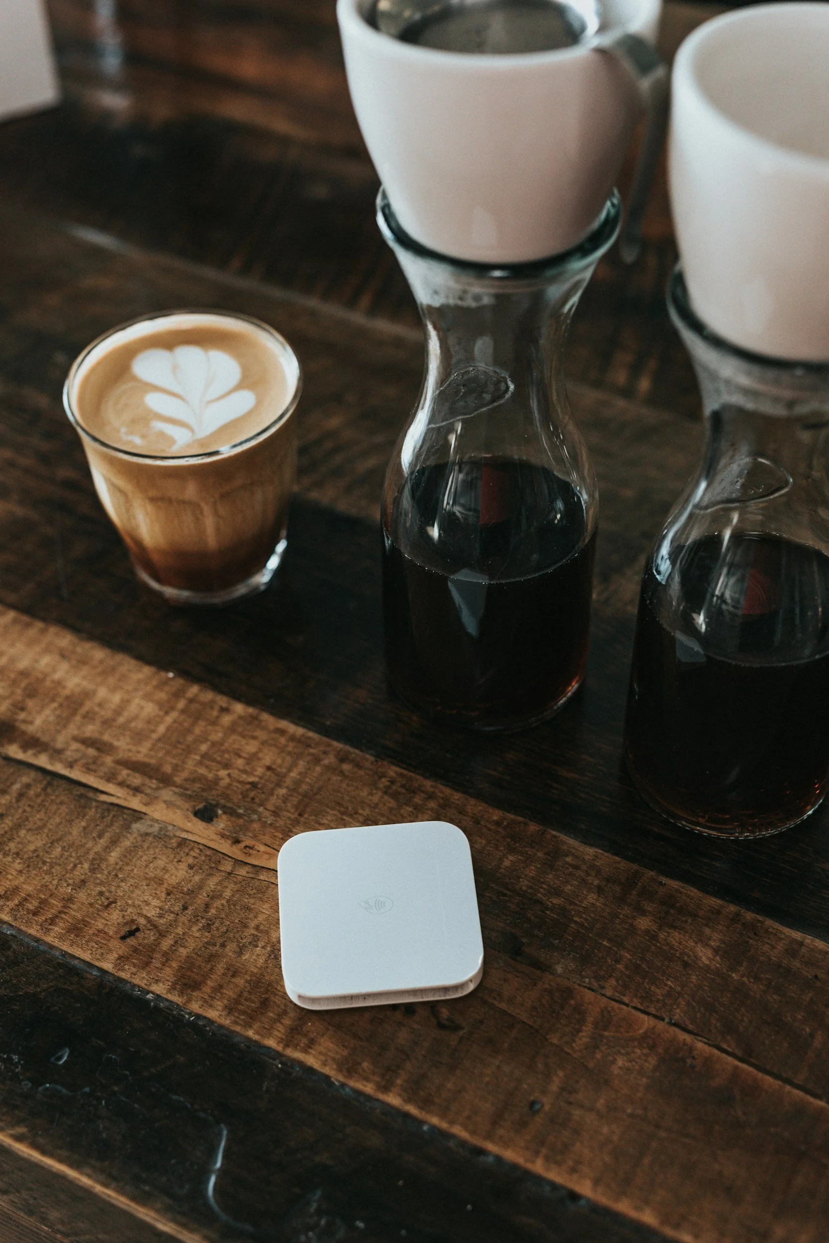 Pour-over coffee setup with a latte art coffee, three carafes of dark coffee, and a wireless device on a wooden table.