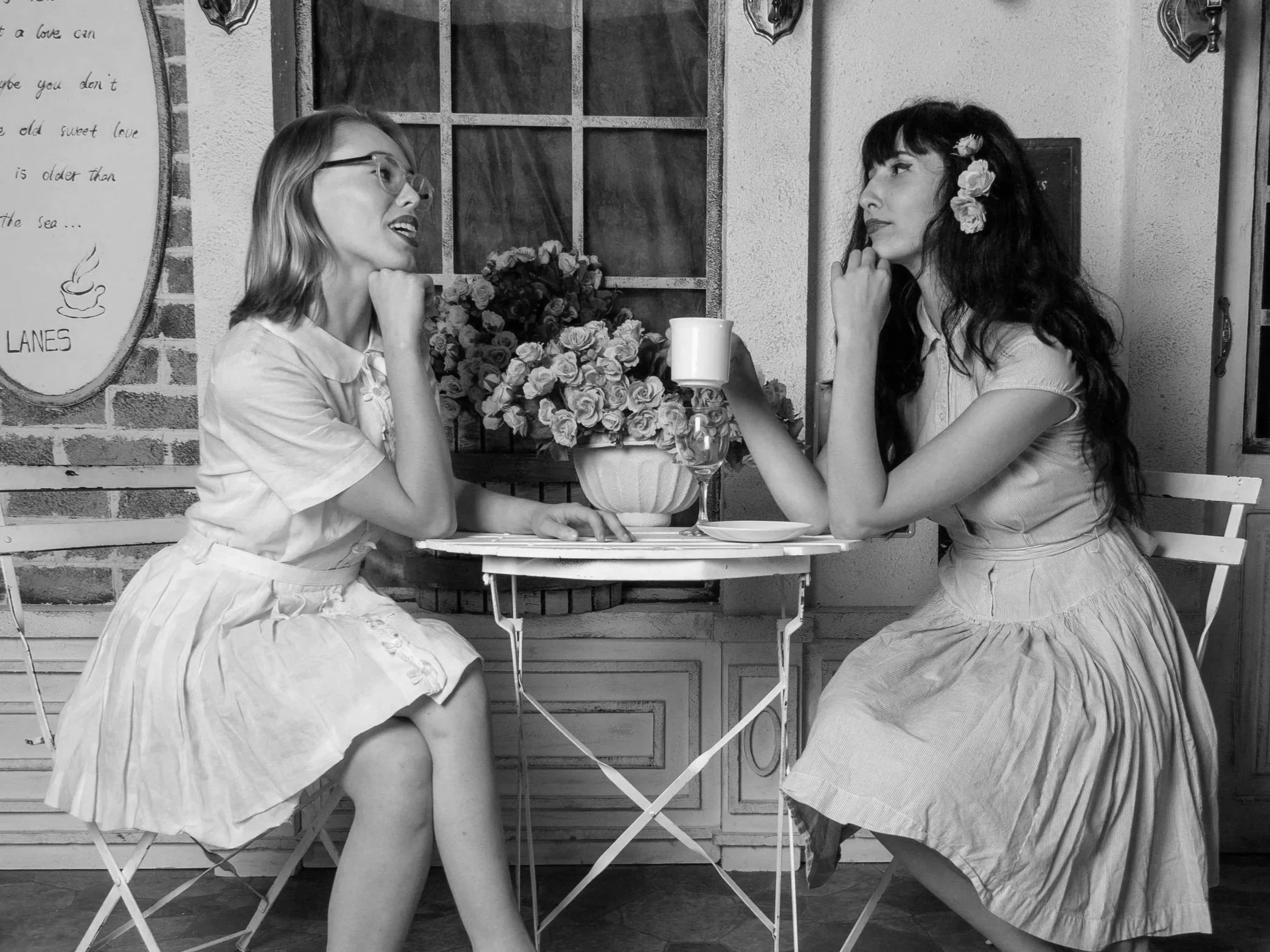 Two women sitting at a small round table in a café, engaged in conversation. One woman holds a cup, and the other woman is smiling. There is a vase with flowers on the table, and a menu or sign on the wall behind them.