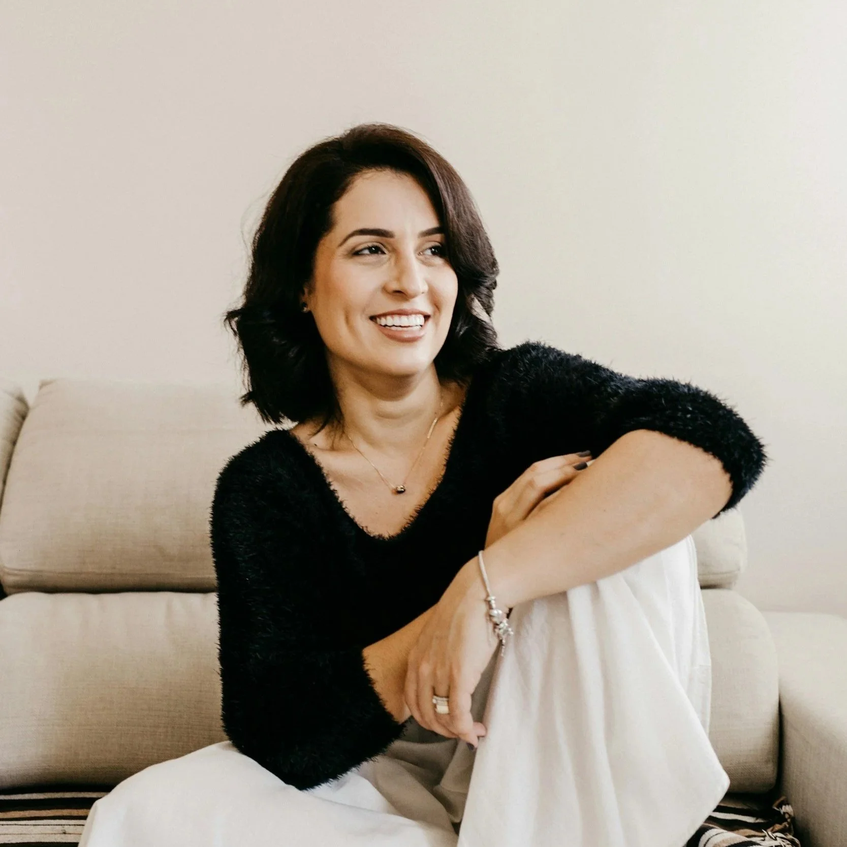 A woman with dark hair, smiling, sitting on a beige couch in a casual pose, wearing a black sweater and white pants.