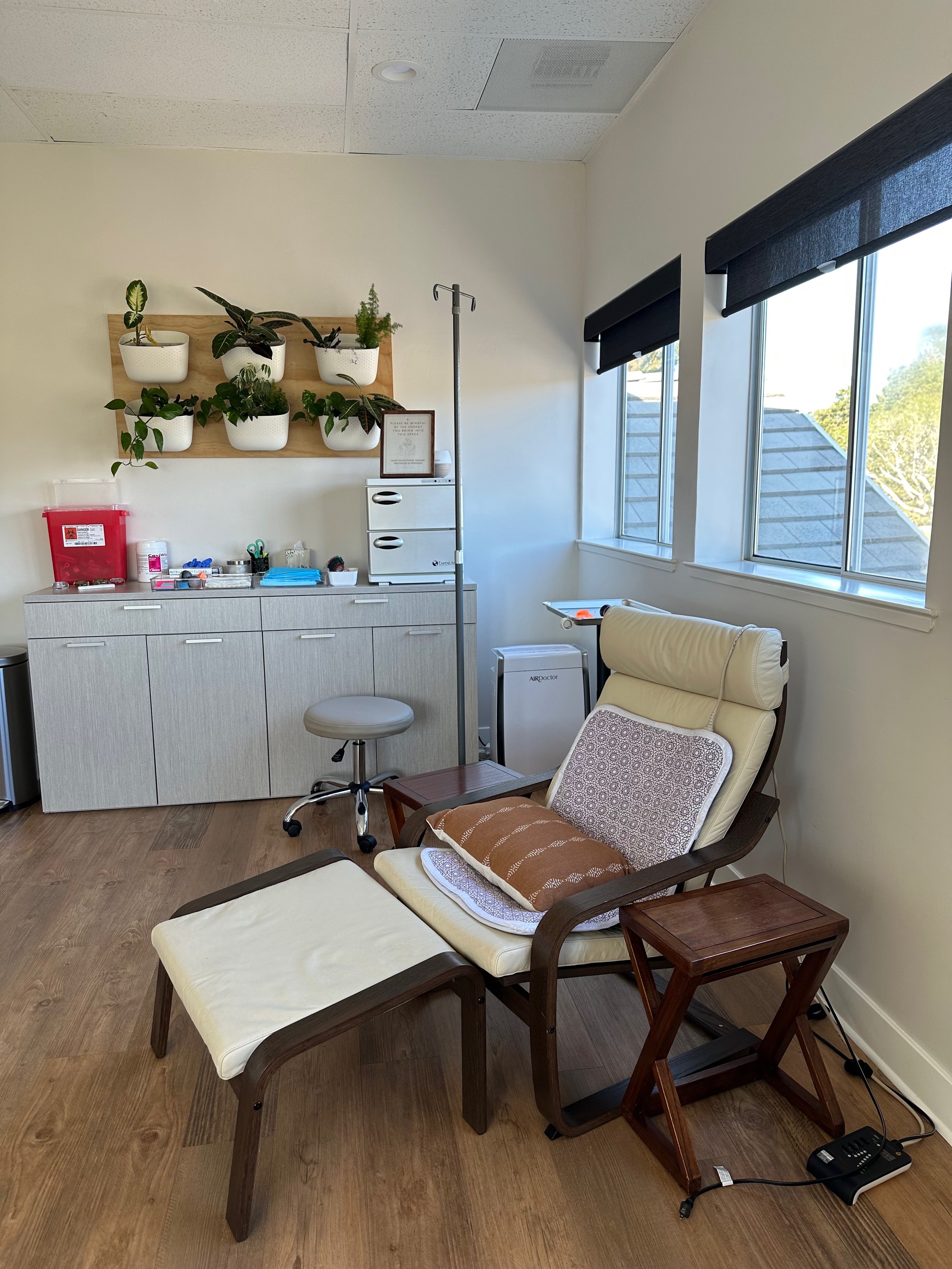 Medical office room with a hospital bed, cushioned chair, plants on the wall, IV pole, and a cabinet with medical supplies.