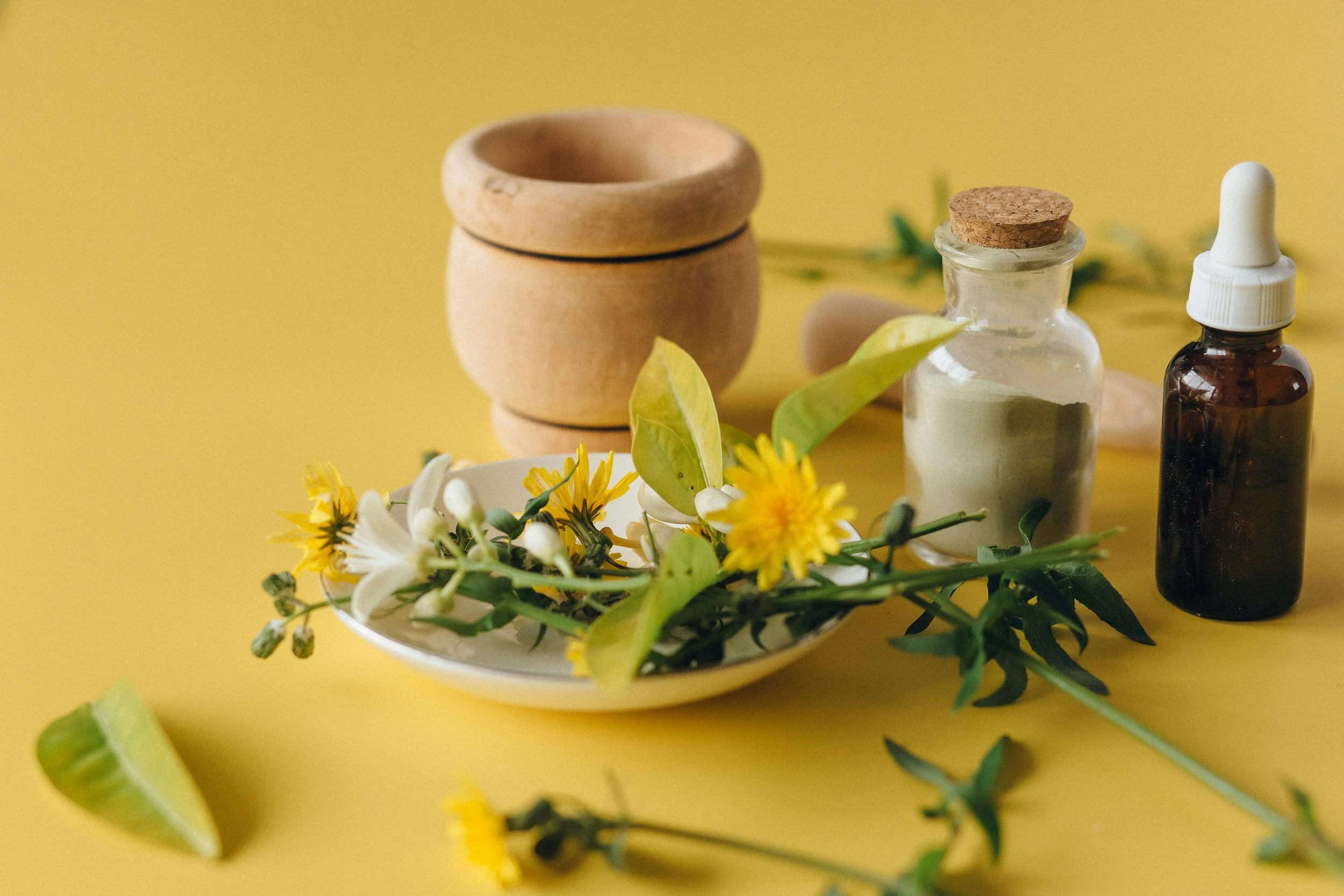 A yellow background with a small white dish holding yellow and white flowers, green leaves, and a lime wedge. In the background, there are wooden bowls, a small glass jar with a green powder and a cork lid, and a small amber glass bottle with a white dropper cap.