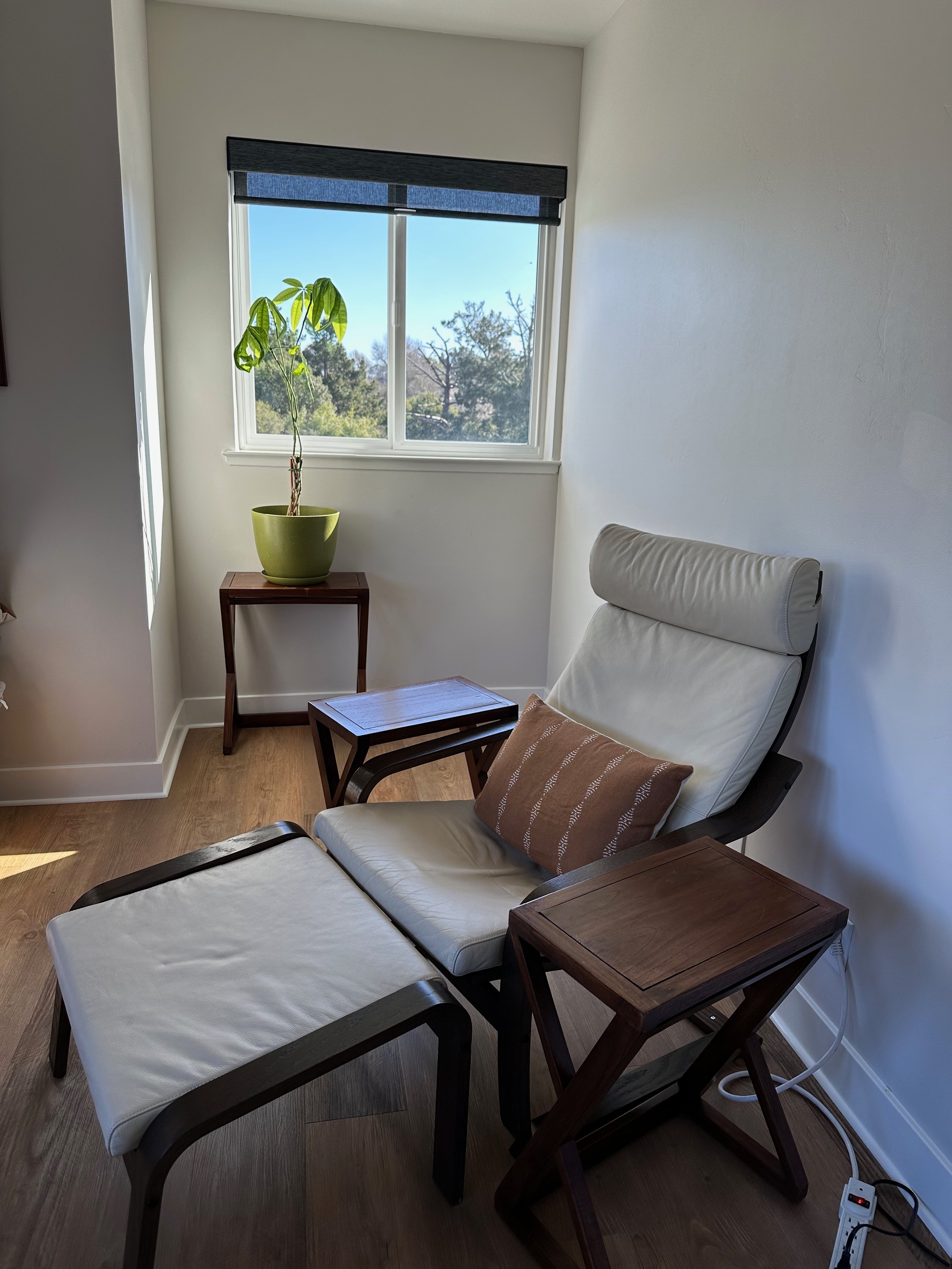 A cozy corner with a white recliner chair, an ottoman, and two wooden side tables next to a window with a view of trees and a blue sky. A potted plant is on a small table by the window, with sunlight streaming in.
