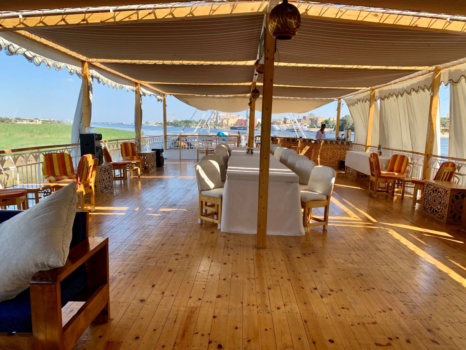 Erholsamer Blick auf eine bewohnte Flussseite mit einem hell erleuchteten, offenen Holzdeck, dekorativen Möbeln und einem Barbereich im Freien bei Tageslicht.