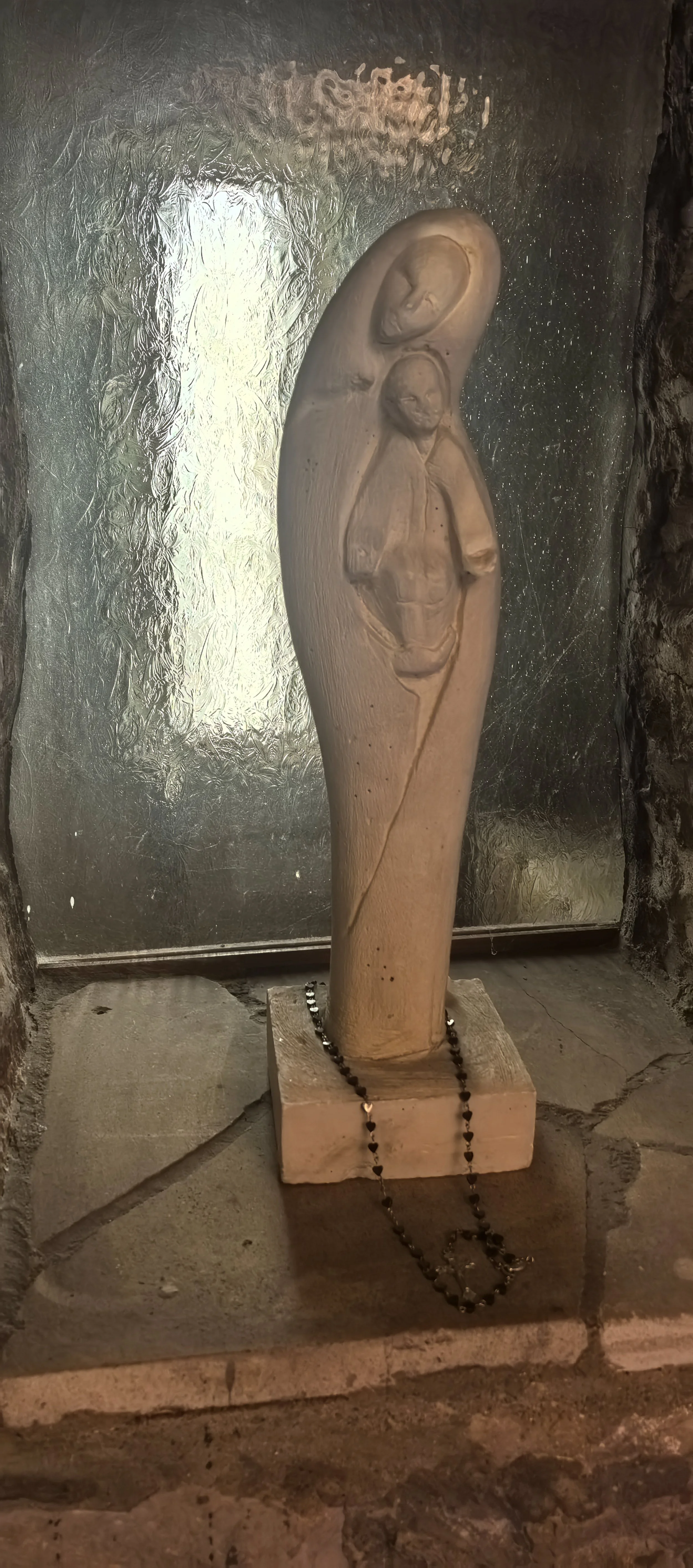 Skulptur eines menschlichen Gesichts, das an einem Holzblock befestigt ist, mit einer schwarzen Perlenkette davor, vor einem metallischen Hintergrund.