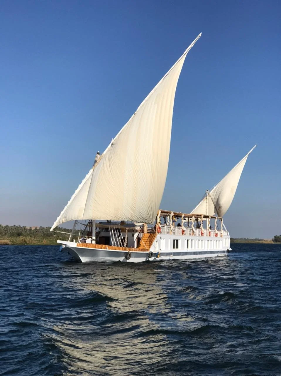 Ein Segelboot mit weißen Segeln auf dem Wasser, im Hintergrund eine Uferlandschaft bei Sonnenuntergang.