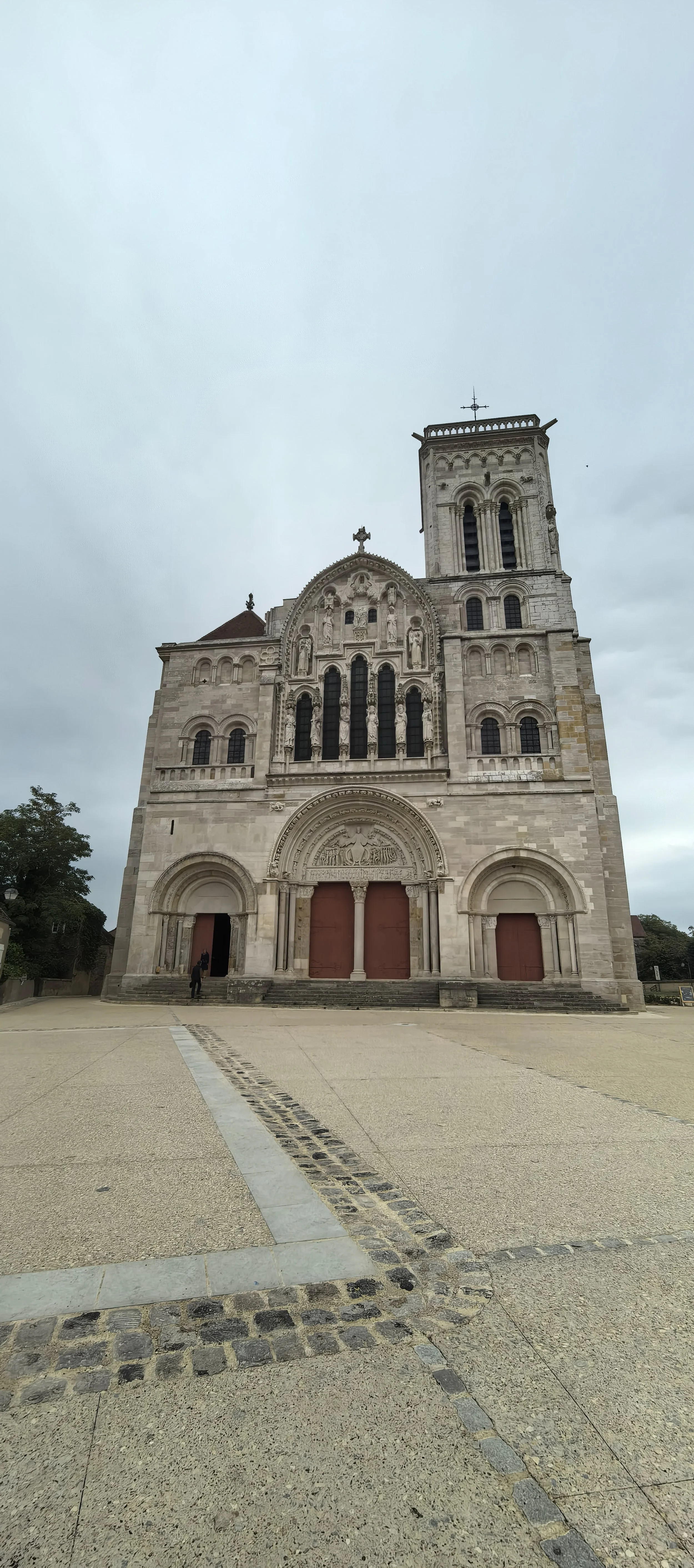 Ein mittelalterliches Kirchengebäude mit einem großen Turm, steinernen Fassaden, großen Türen und kunstvollen Skulpturen über dem Eingang, vor einem steinernen Platz mit einem Fußweg.