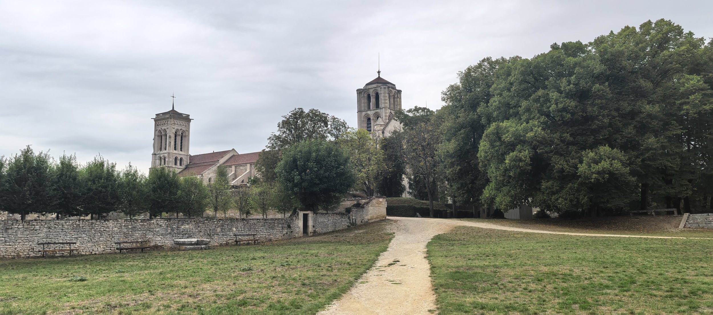 Eine ländliche Szene mit einem Weg, Bäumen, einer Steinmauer und einer Kirche im Hintergrund, die bei bedecktem Himmel fotografiert wurde.