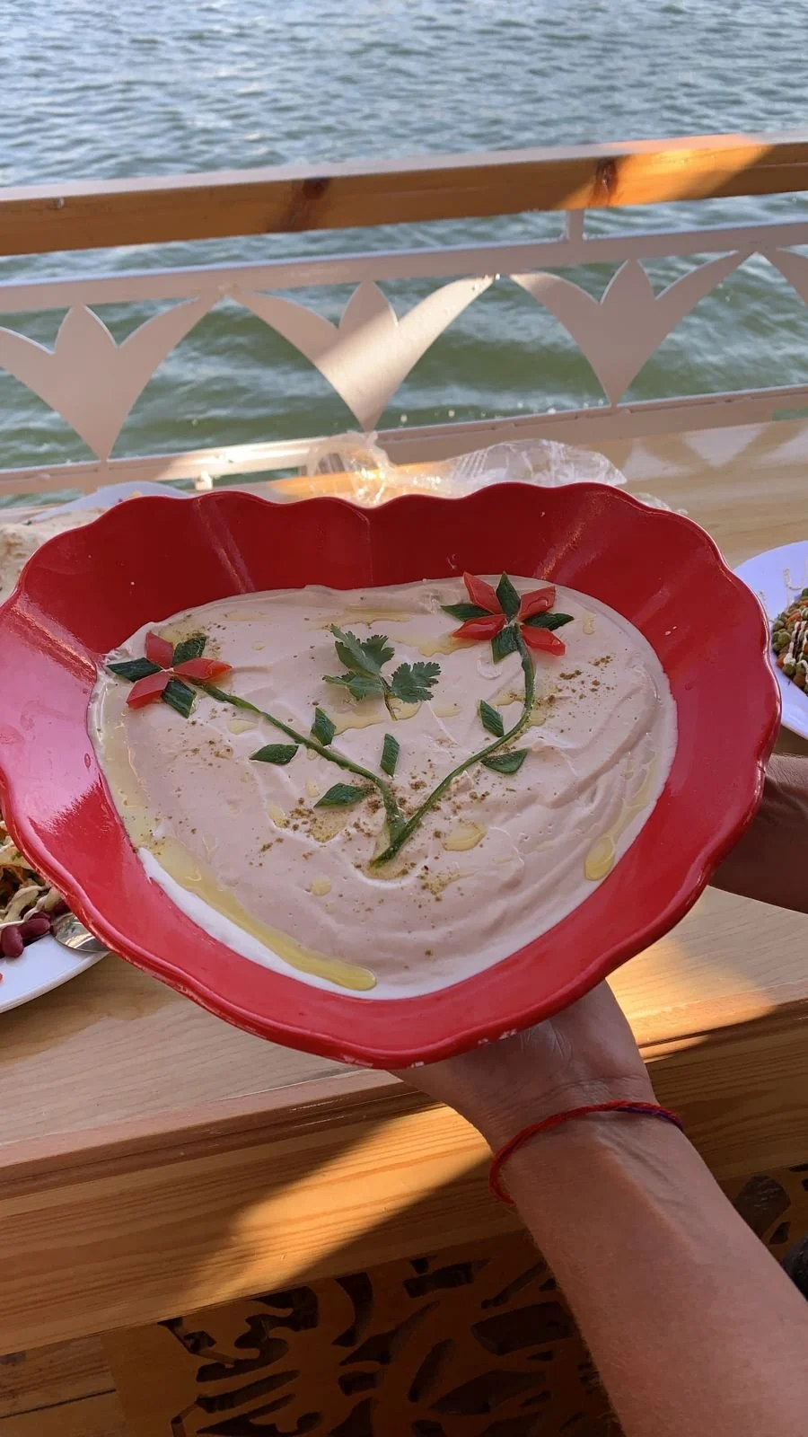 Eine rote Schale mit einem cremigen Dip, dekoriert mit kleinen roten und grünen Gemüseschnitzereien und frischer Petersilie, am Wasser mit einer Holzbrücke im Hintergrund.