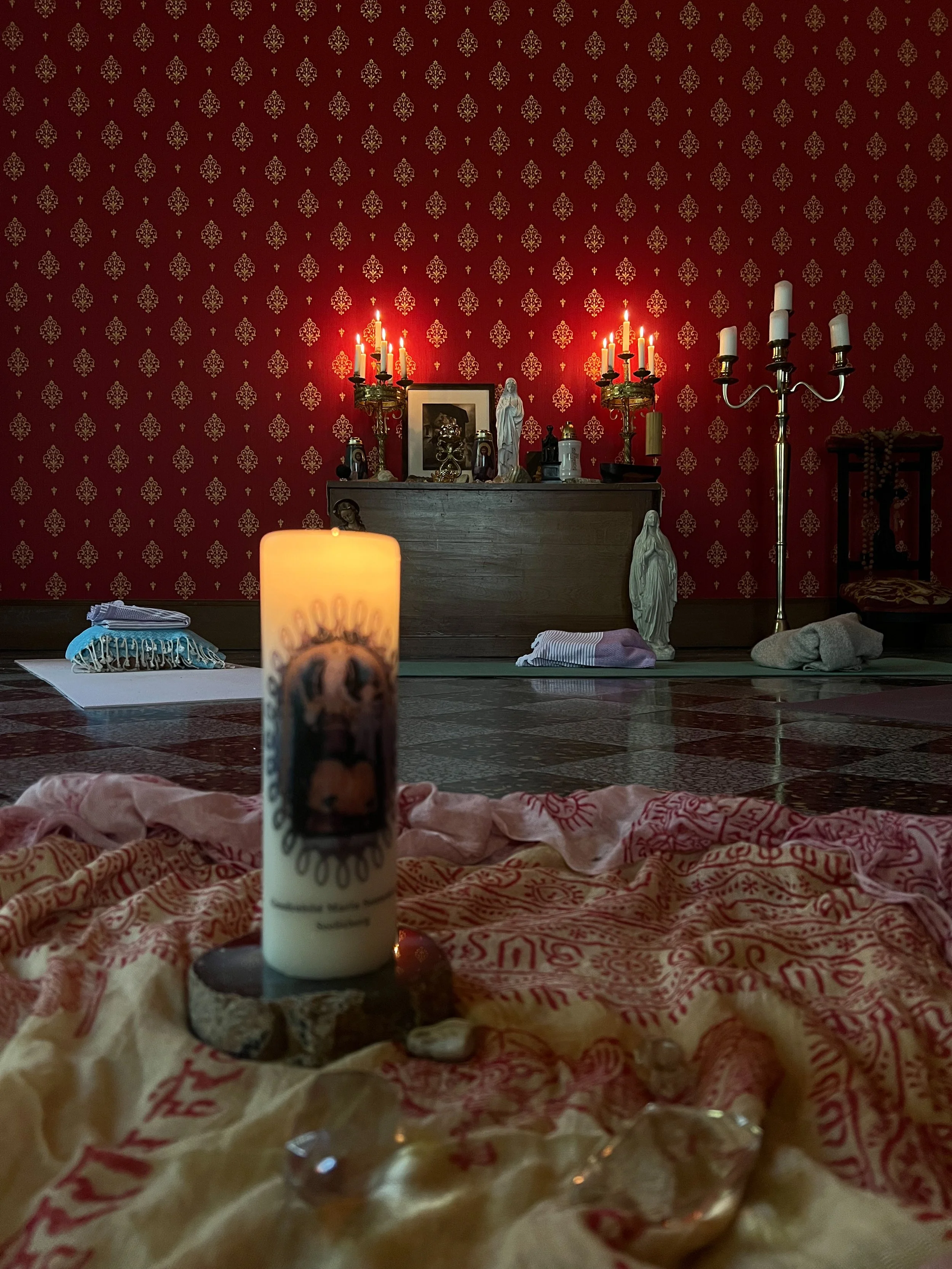 Ein Altar mit Kerzen, Figuren und religiösen Gegenständen vor einer roten Wand mit goldnem Mustermotiv in einem Raum.