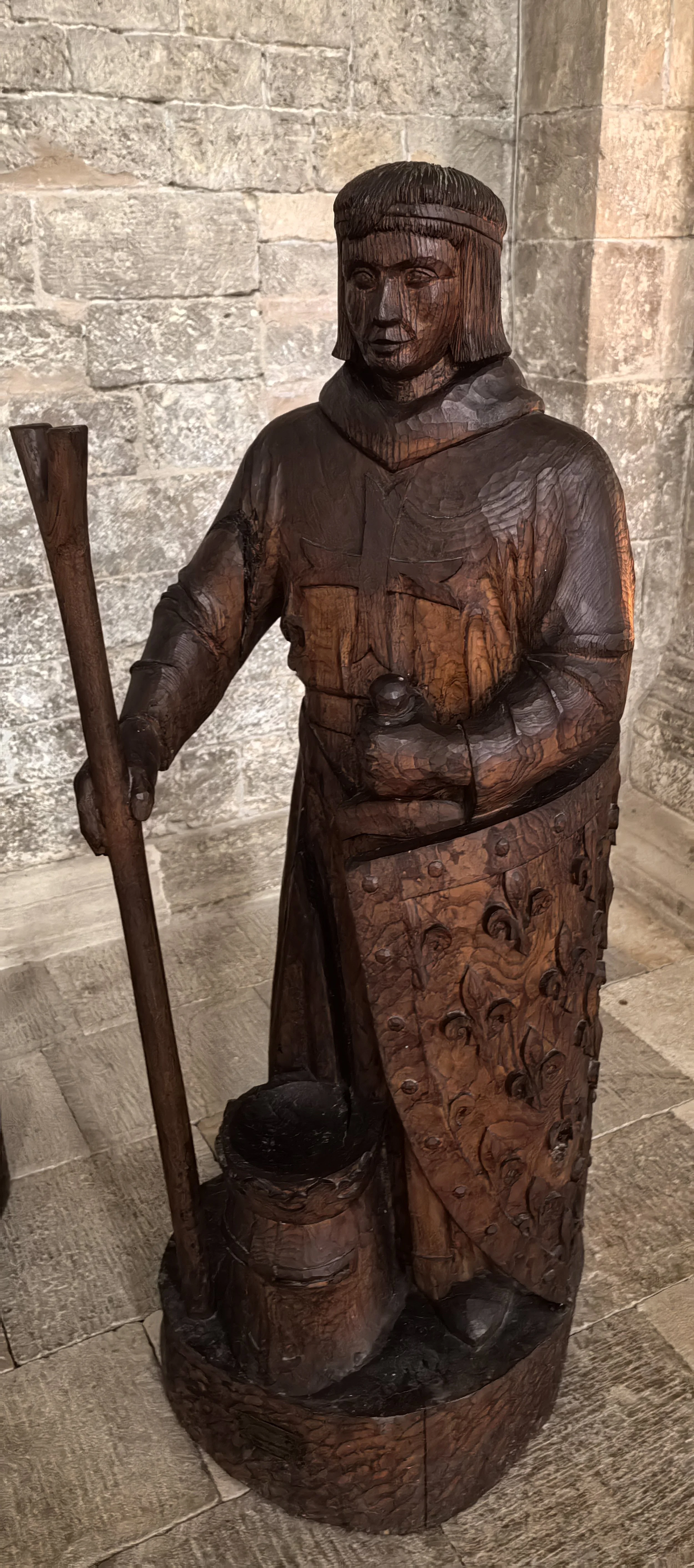 Eine Holzschnitzfigur eines Mädchens in historischem Kleidung mit einem Eimer und einem Stock, vor einer Steinmauer.