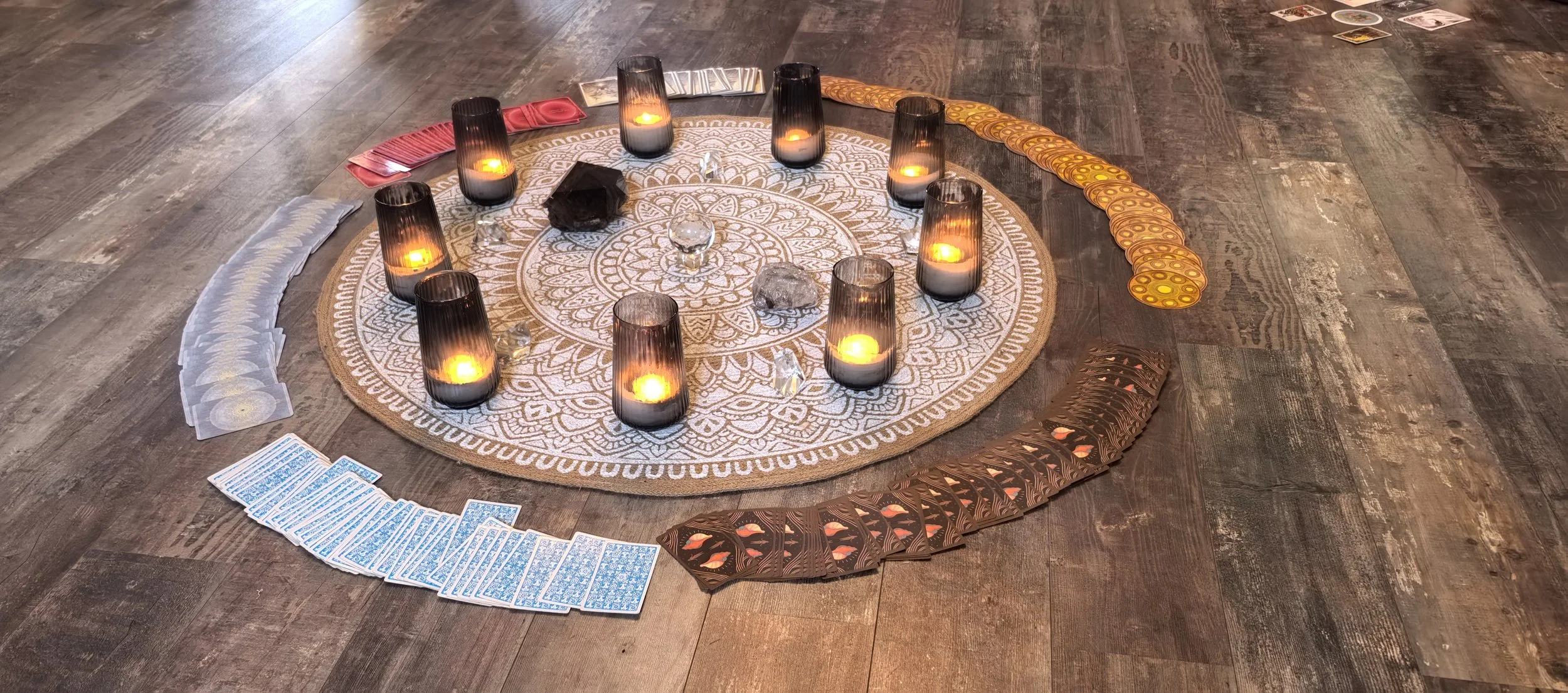 Rund um einen weißen Mandala-Teppich liegen Tarotkarten, Kristalle und Steine, umgeben von neun schwarzen Kerzen in Glasgefäßen. Das Setting ist für eine spirituelle oder esoterische Praxis vorbereitet.