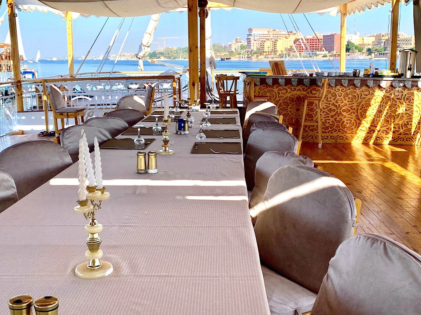 Ein festlich gedeckter Essbereich mit einem langen Tisch, Stühlen, Kerzen und Besteck, mit Blick auf das Wasser und Boote im Hintergrund, im Freien unter einem Sonnenschirm.